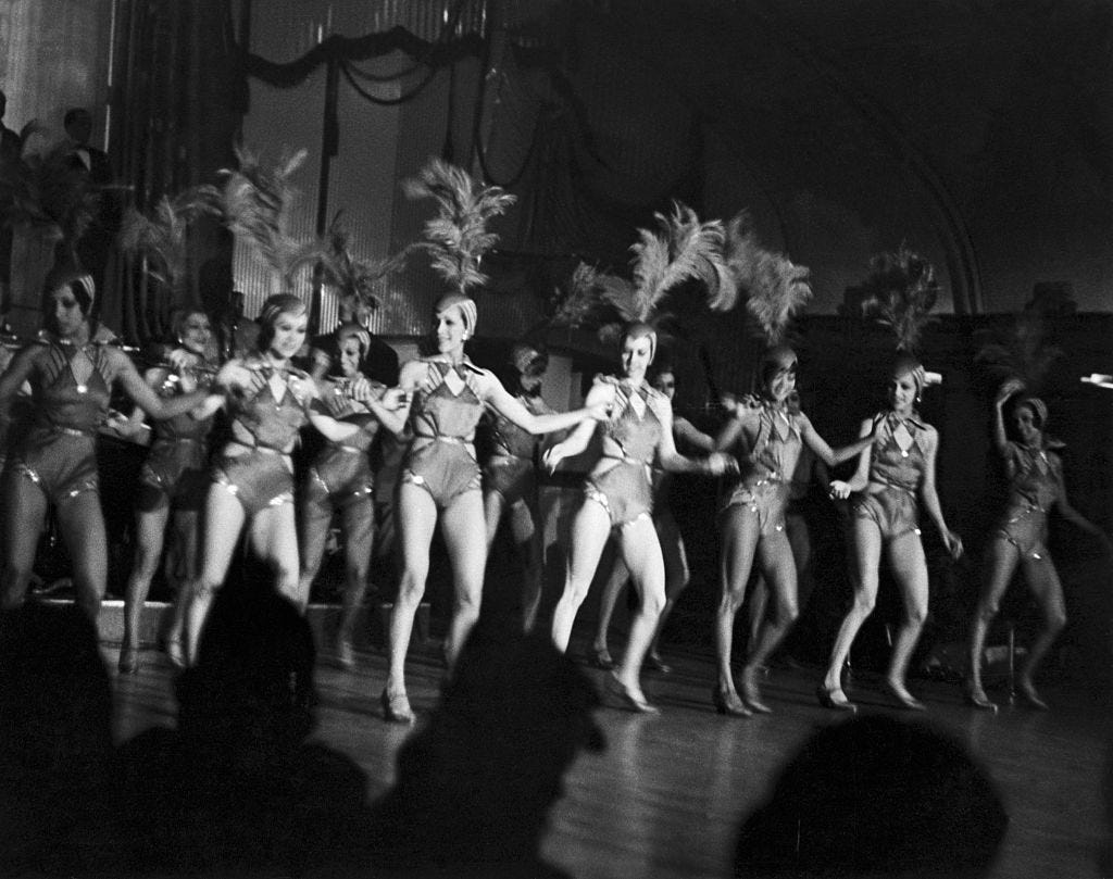A row of chorus girls holding hands and dancing. They're wearing feathered hats and pointed stilettos.