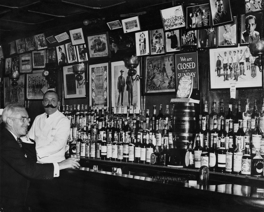 A well-stocked bar at Bill's Gay Nineties, a speakeasy located at 57 East 54th Street, New York City. The barman is wearing a large fake handlebar mustache.