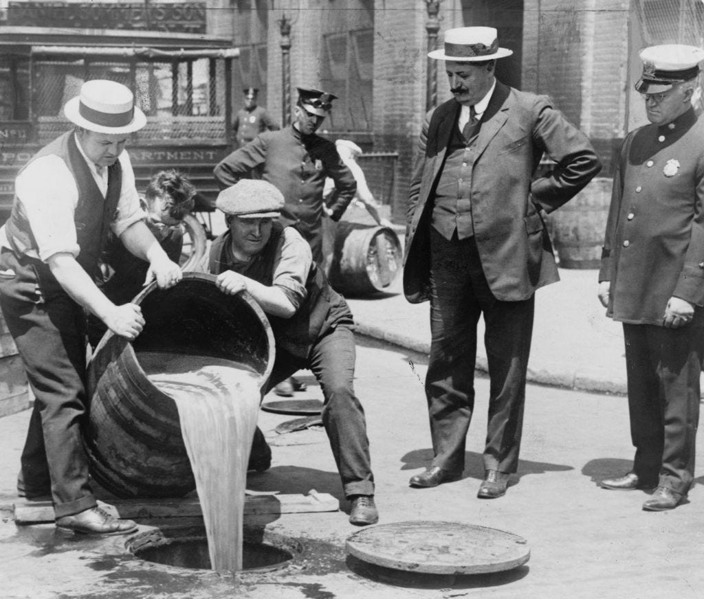 Three policemen watch on as two men pour a barrel filled with alcohol down the drain.