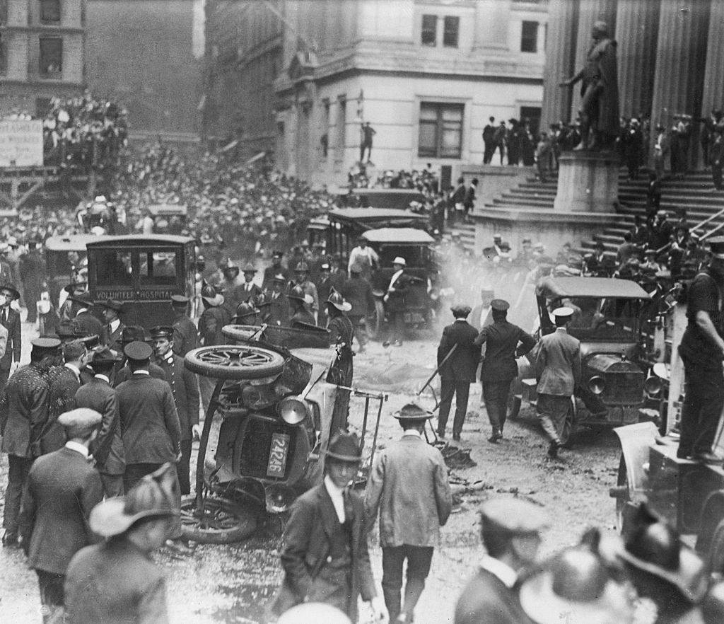 Several people crowd around a car outside Wall Street. The car was bombed, and parts of it lay around on the road.