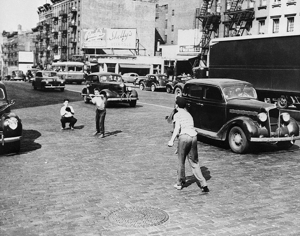 Three boys playing stickball on a busy street in Manhattan, surrounded by cars.