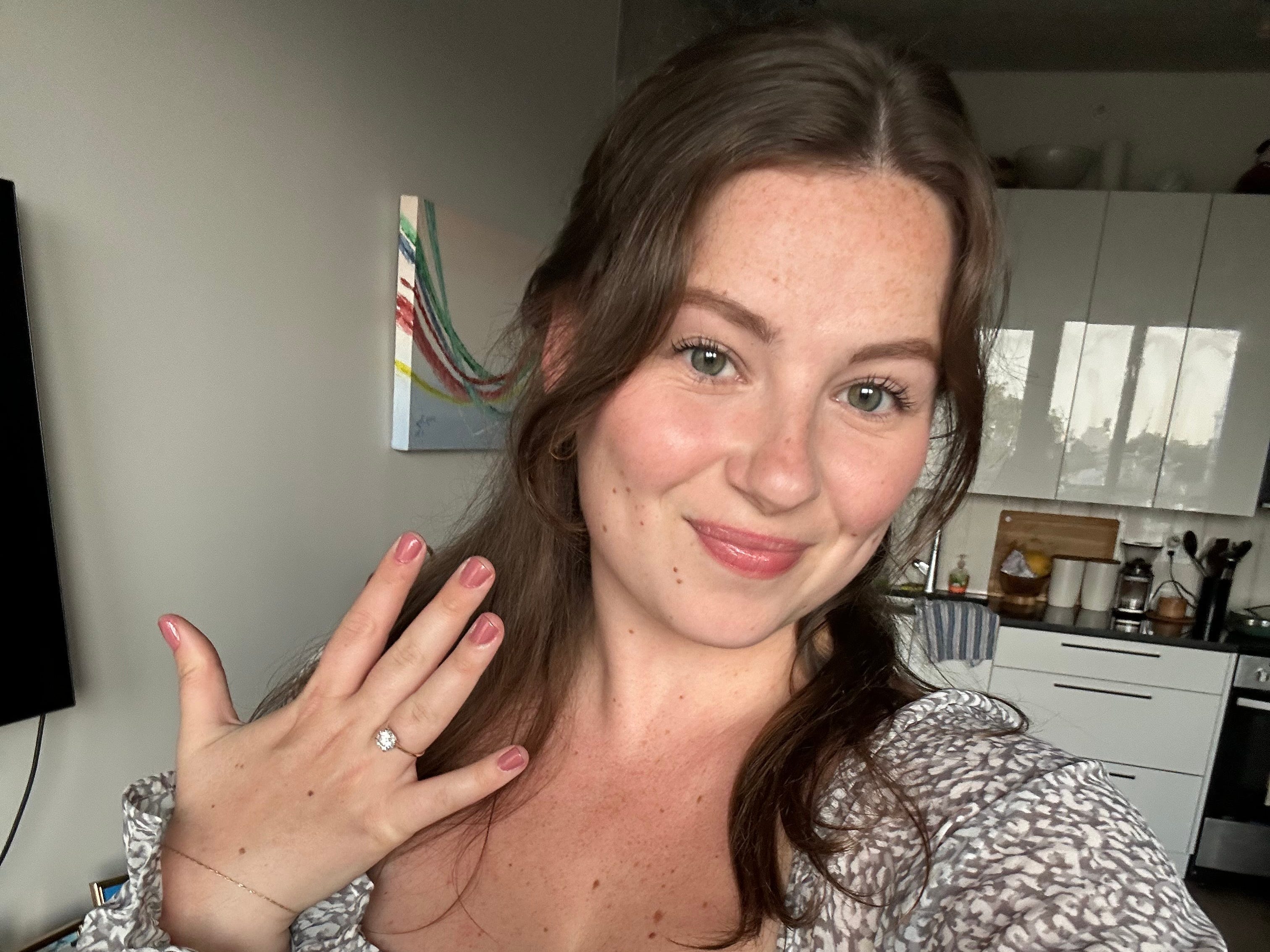 A woman stands in a kitchen and holds up er engagement ring.