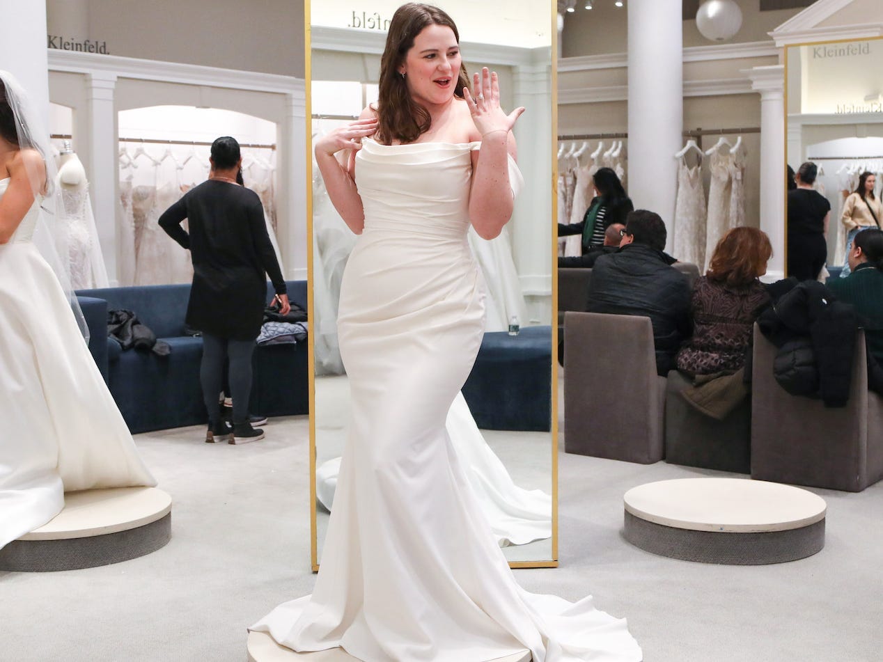 A woman holds up her ring finger while she wears a wedding dress in a bridal boutique.