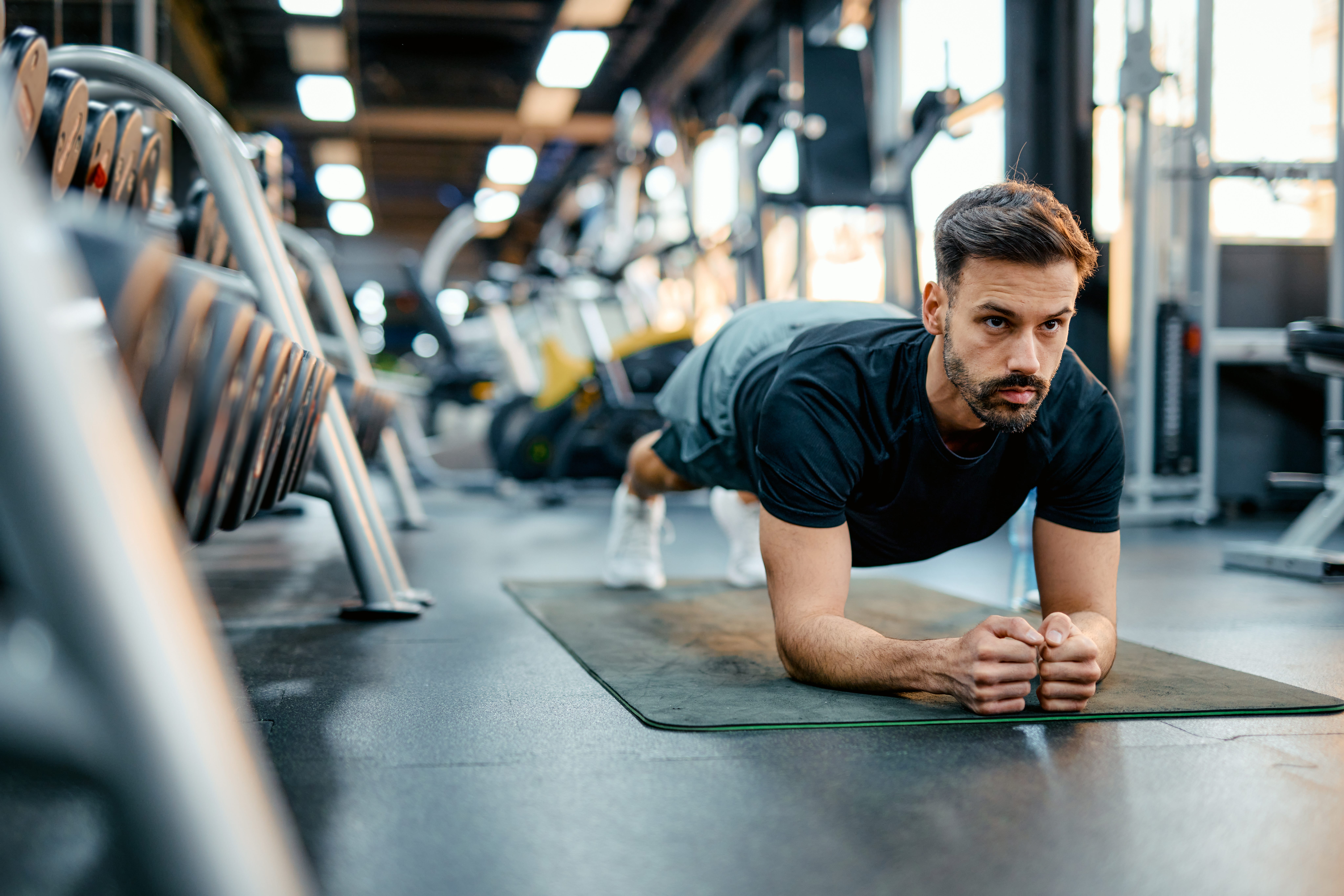 An athletic man doing a plank at an indoor gym with lots of machines.