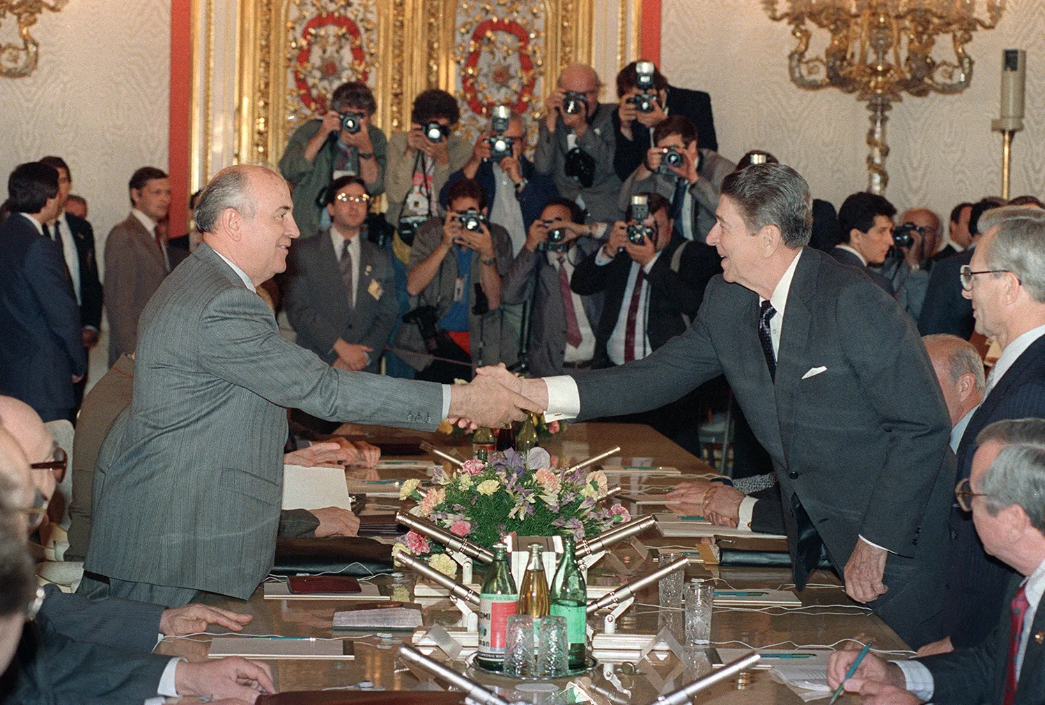 Gorbachev stands and reaches across a table to shake hands with Reagan. Both men wear suit. Behind them is an ornate wall and several photographers all pointing their cameras at the two men.