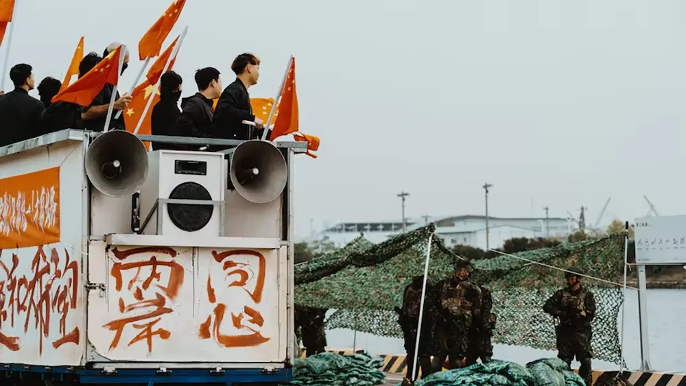 People on a Chinese-flagged vehicle with loud speakers. Below, soldiers on the ground under camo netting.
