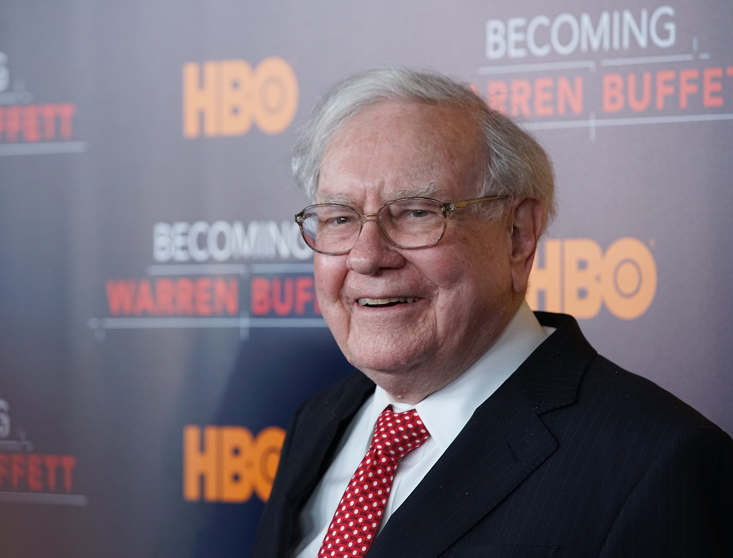 Warren Buffett smiling in a suit standing behind posters for HBO