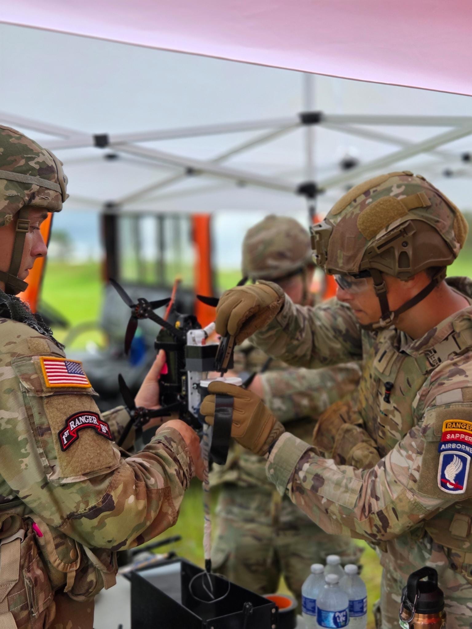 Two US Army soldiers wearing camouflage work on a first-person view quadcopter drone under a tent.