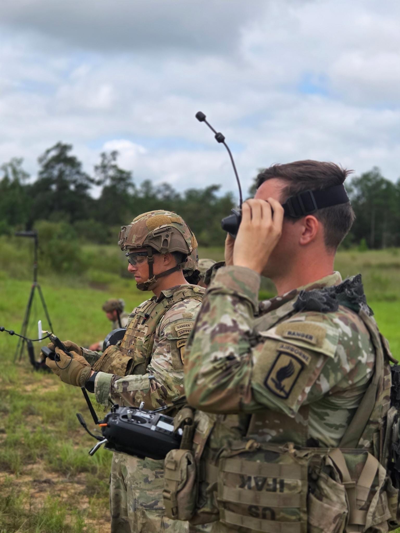 Two US Army soldiers operate drones in a grassy field with a green tree line in the background.