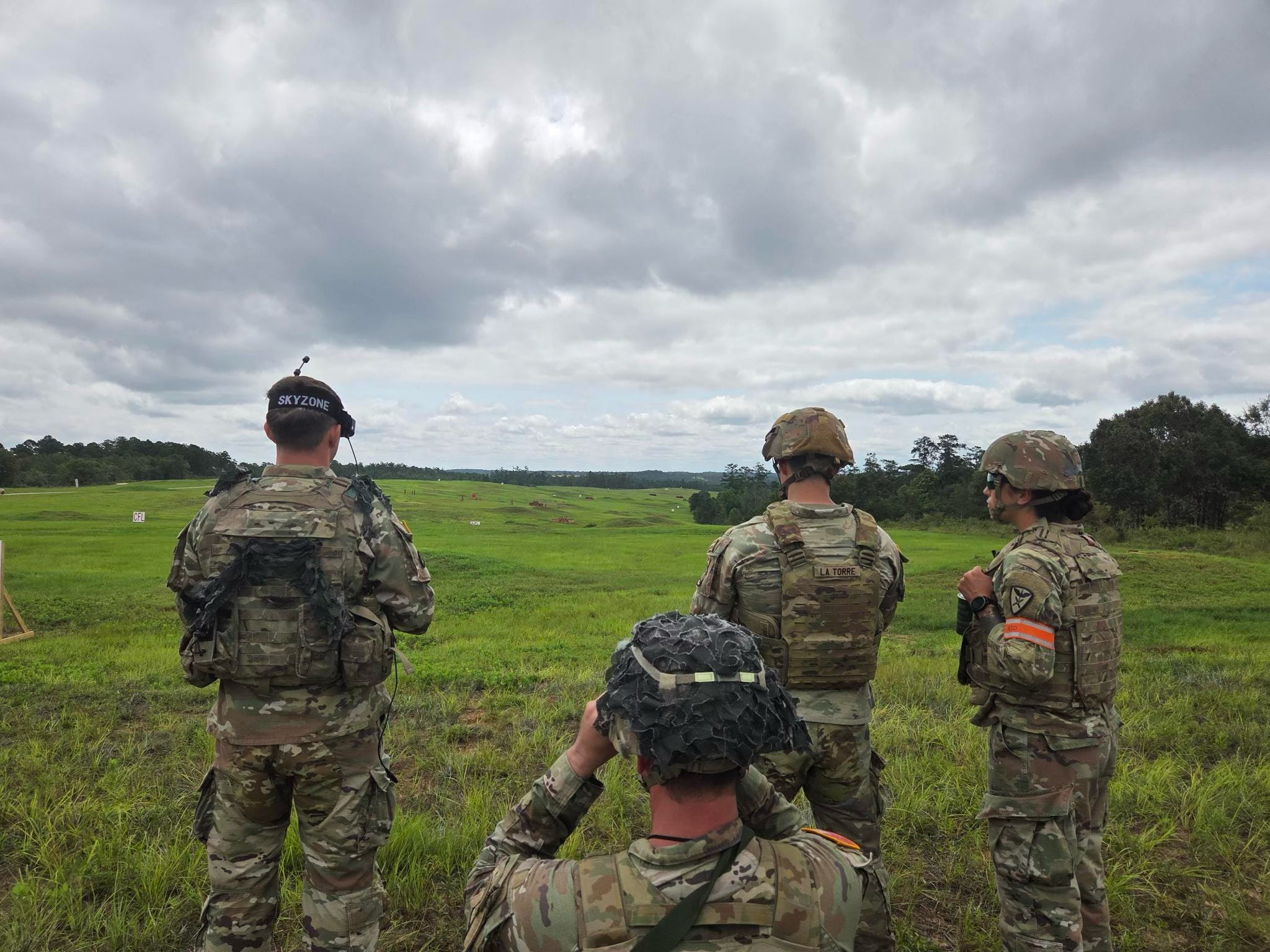 Four US Army soldiers stand in a grassy field looking out to a drone demonstration. the sky is cloudy and grey in the background.
