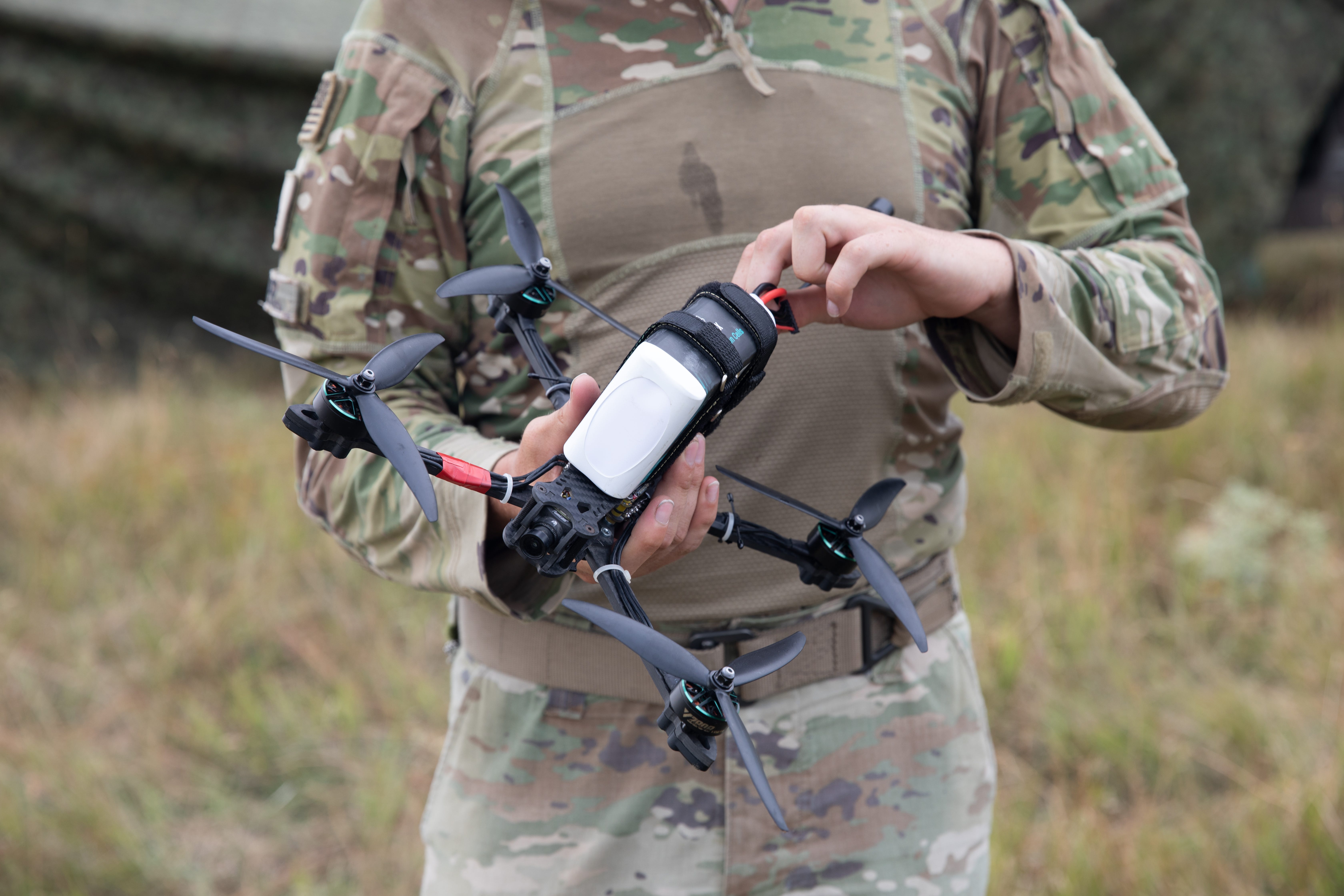 A US Army soldier is seen from the shoulders to the mid-thighs wearing camouflage and holding a quadcopter first-person view drone. The background is greenish brown grass.