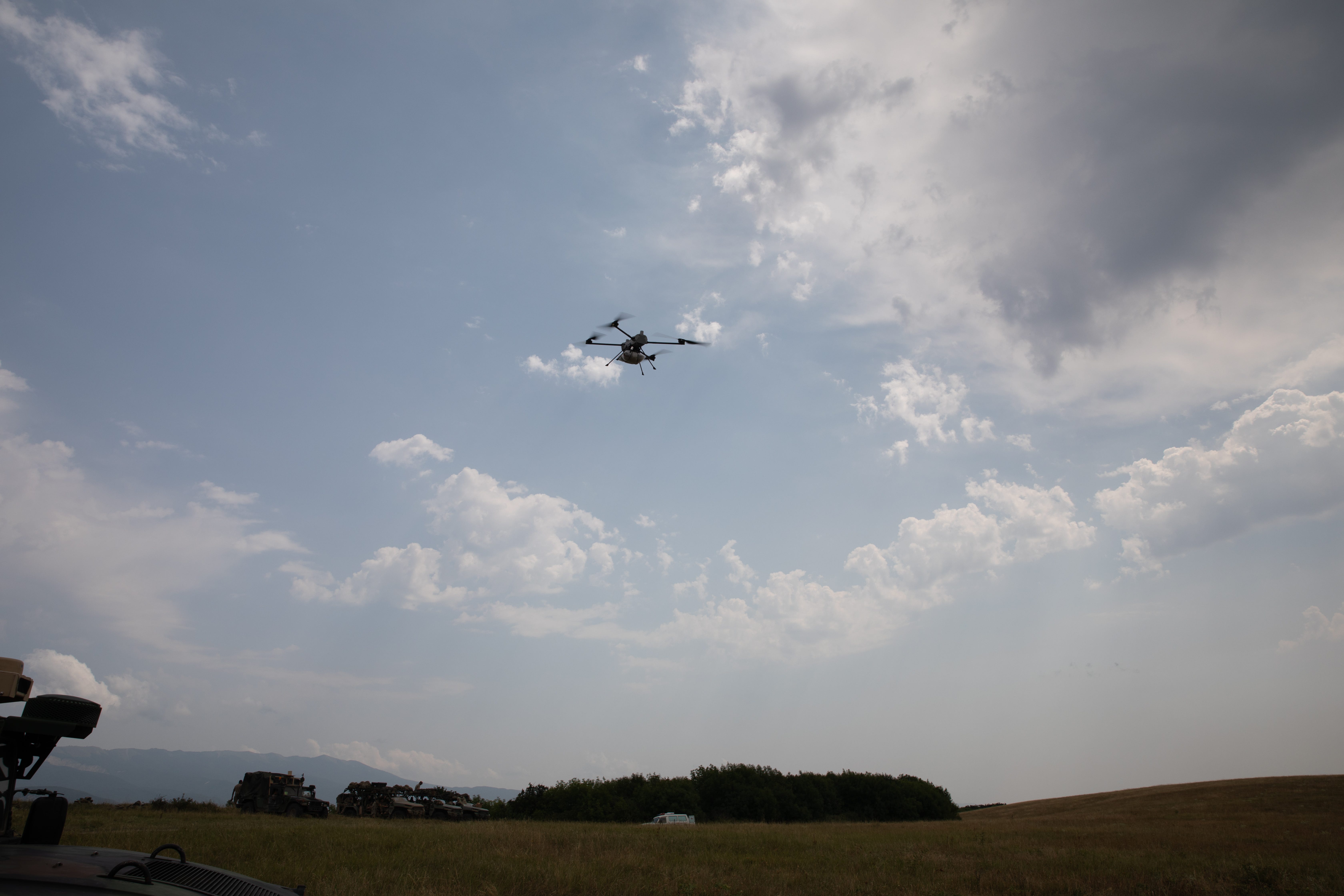 A quadcopter drone flies above a grassy field against a blue and cloudy sky.