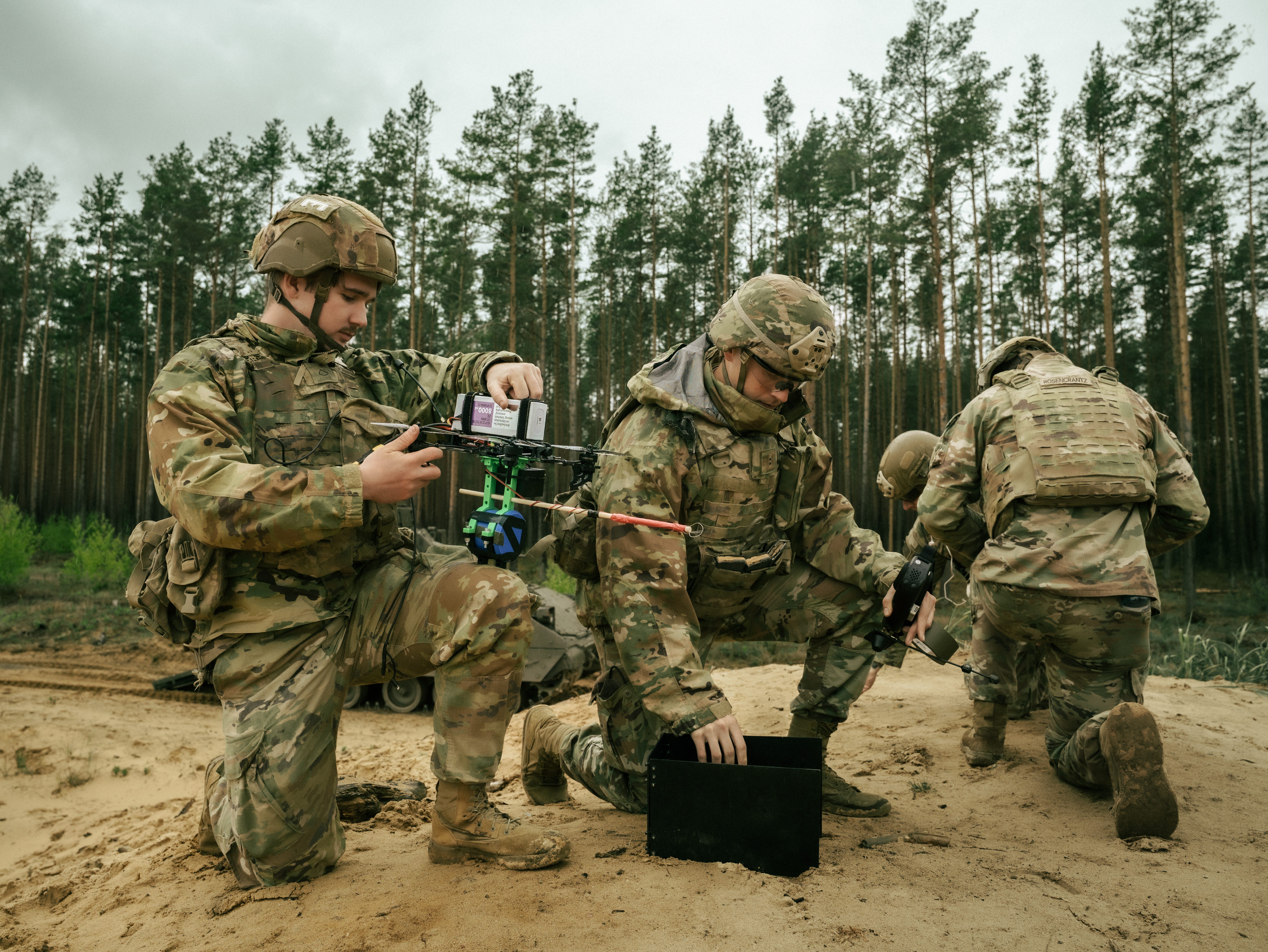 Four US Army soldiers kneel on dirt examining a first-person view quadcopter drone in a forest with a cloudy sky.
