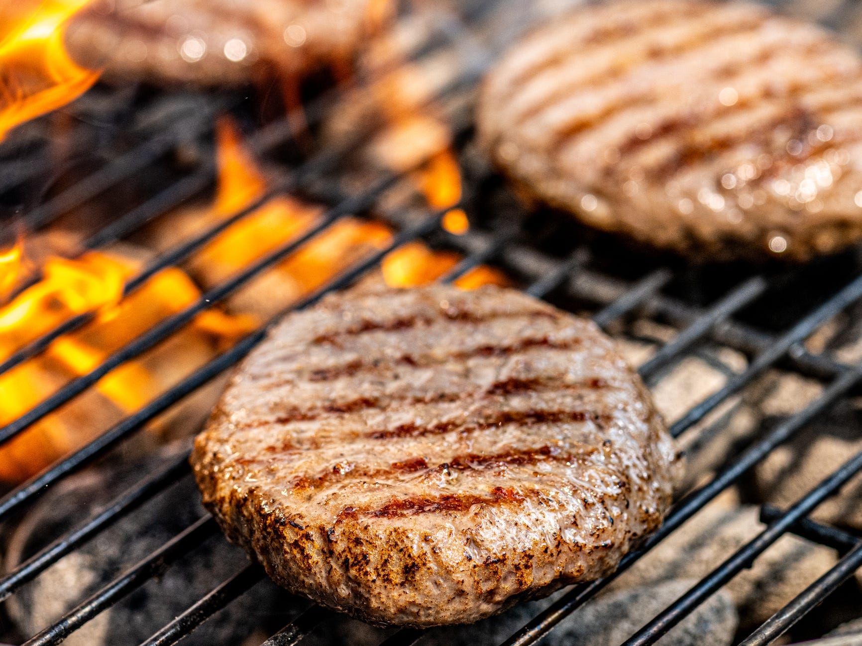 Three brown burgers with grill marks on them sit on a grill as flames rise from the grill in the background