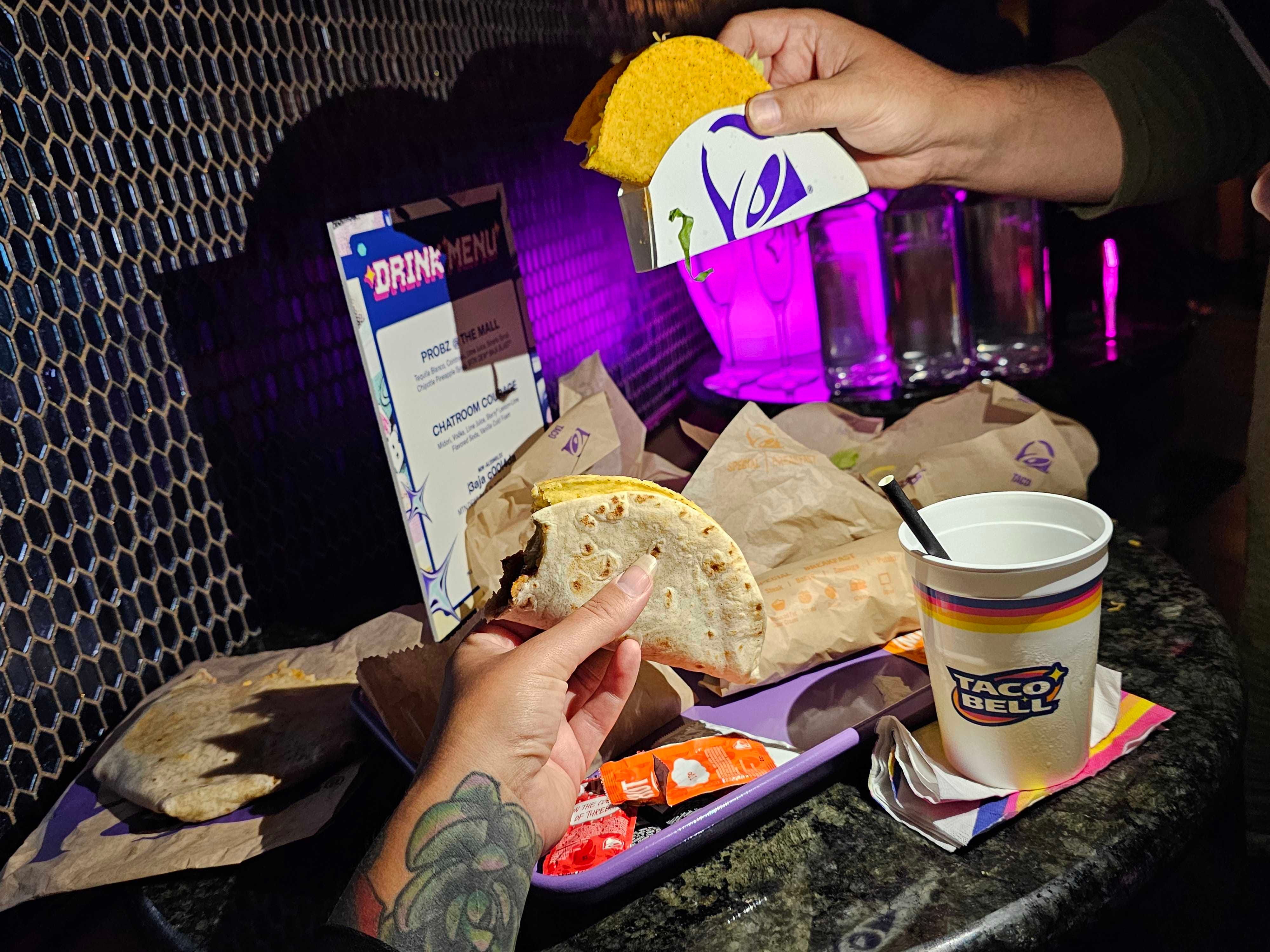 Two hands hold up Y2K-themed Taco Bell products, recently rereleased by the brand: a double decker taco and a cool ranch doritos locos taco.