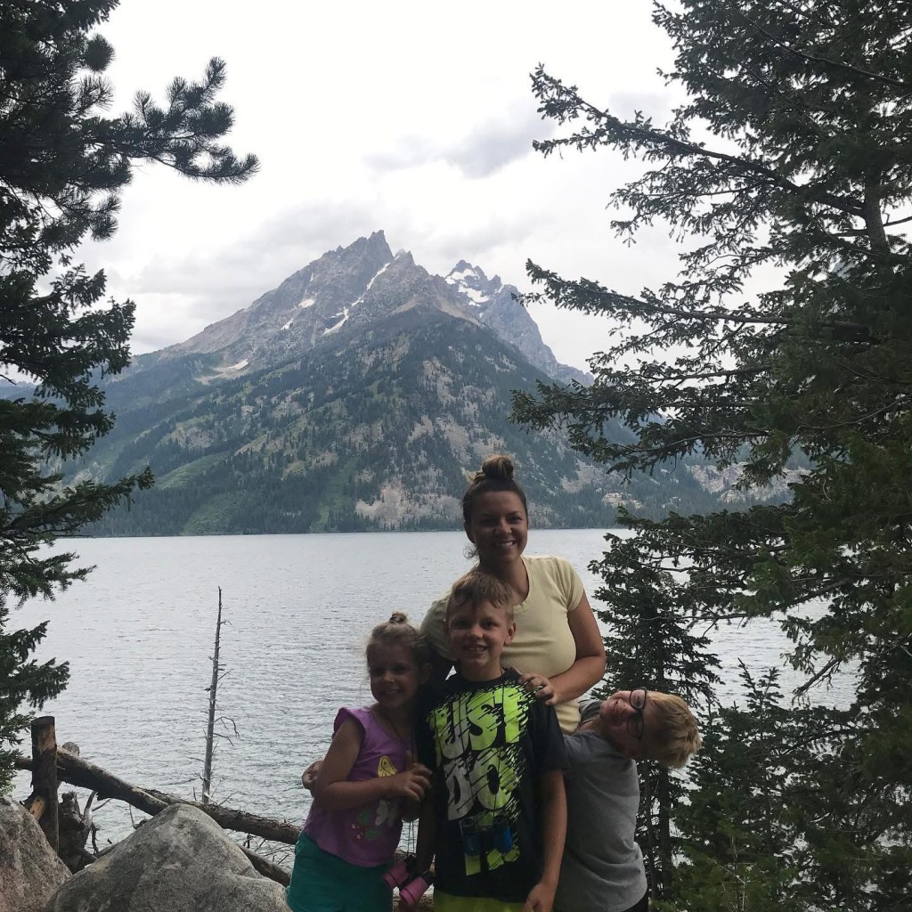 A woman and her three children pose for a photo by a lake with mountains in the background.
