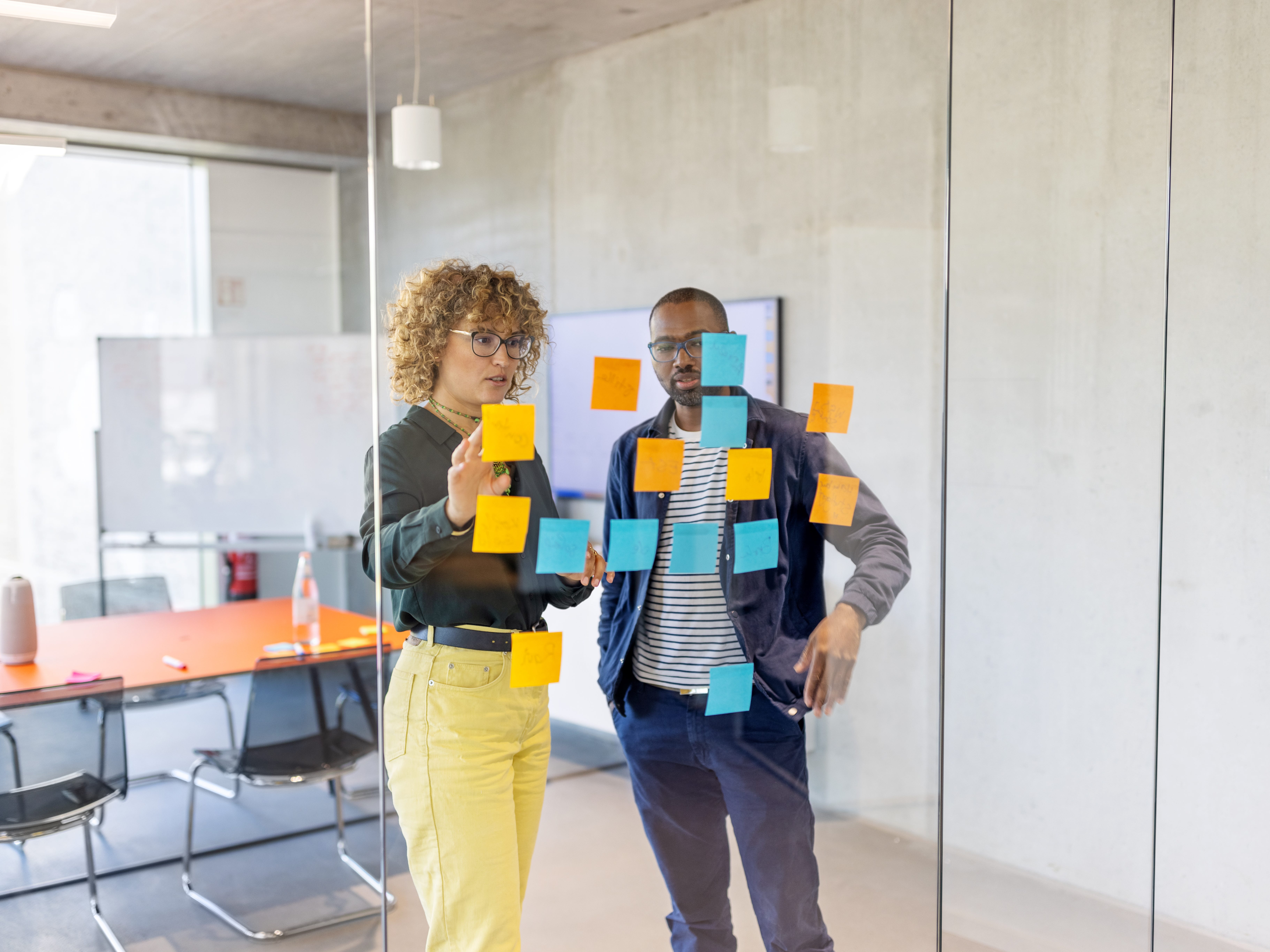 Two people are looking at adhesive notes stuck to a glass wall