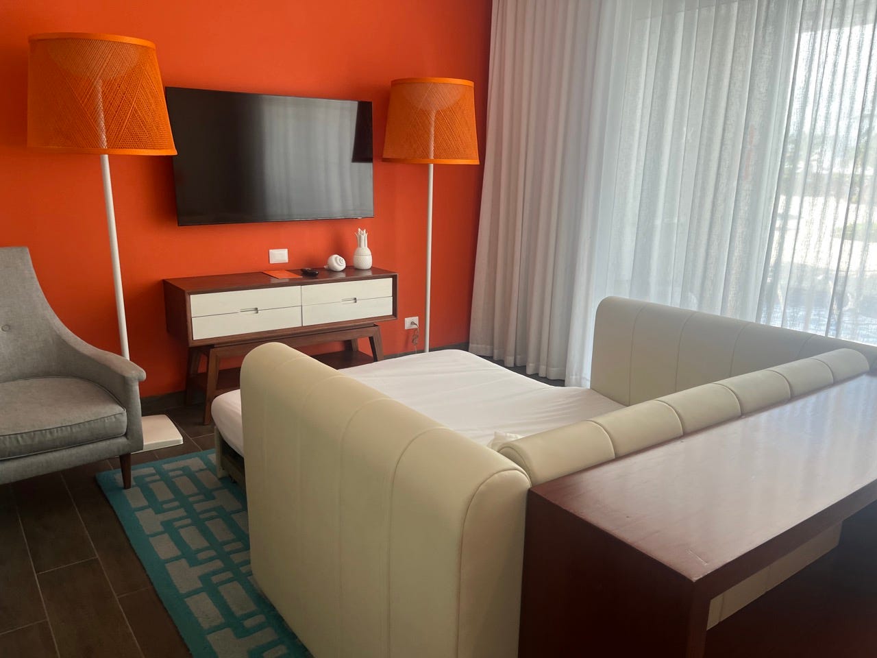 Mounted TV on orange wall with fold-out couch in front of it and orange lampshades on either side of TV