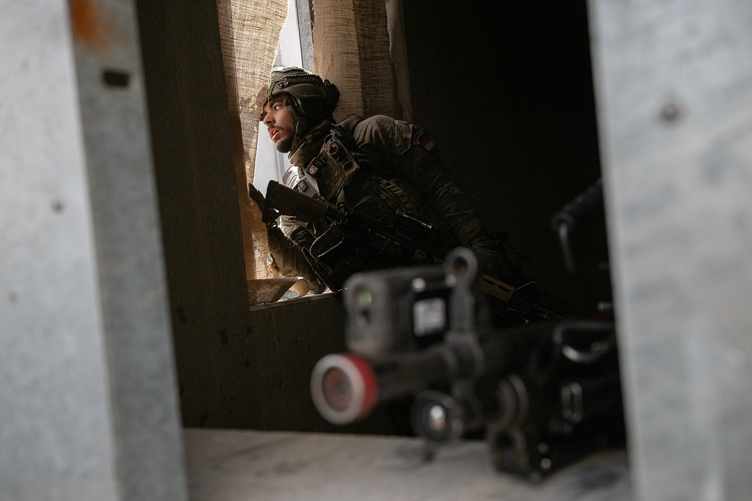 A soldier in full combat gear peers past a curtain from inside a building. A gun with scope is seen on a window in the foreground.