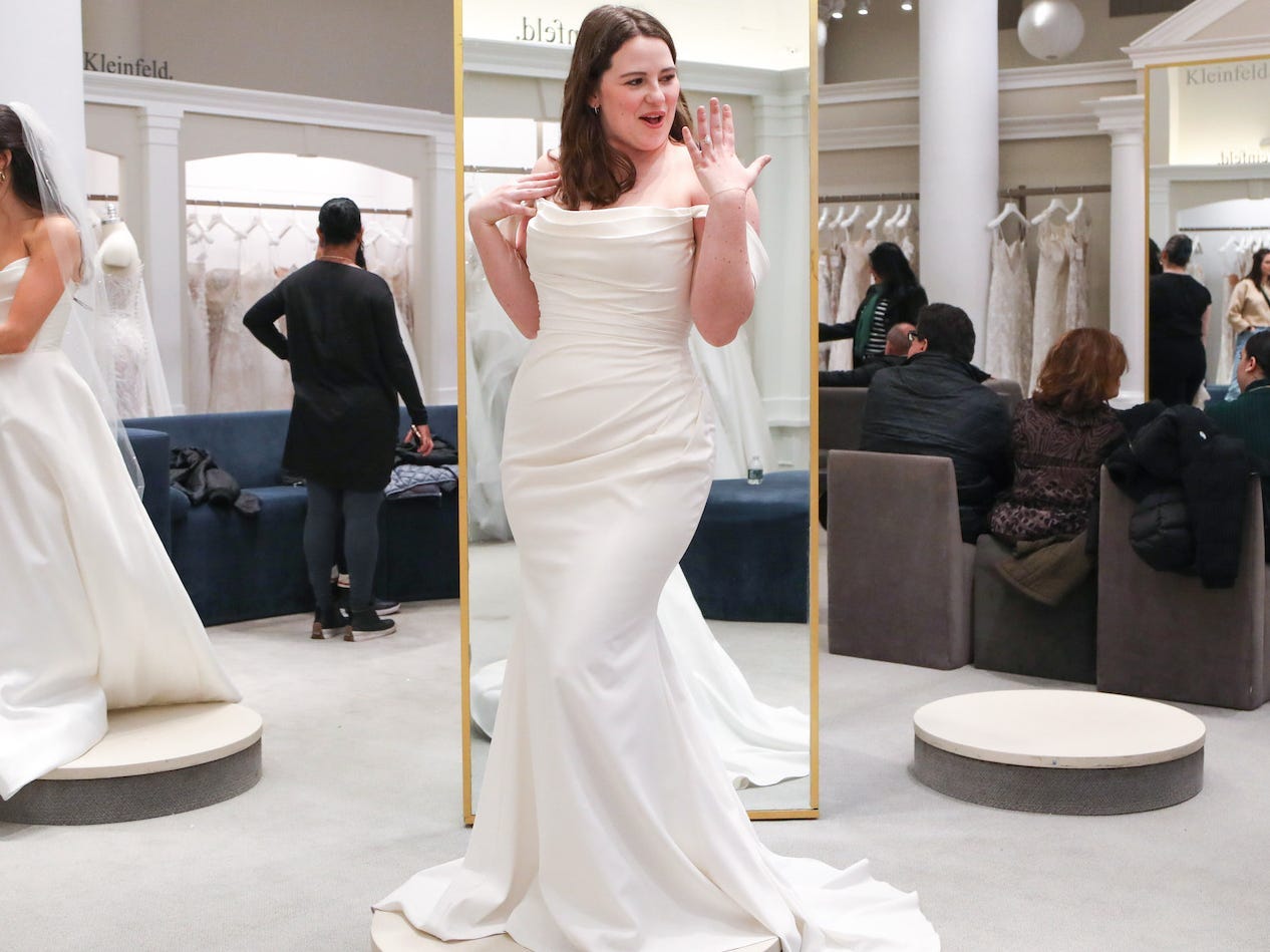 A woman holds up her ring finger while she wears a wedding dress in a bridal boutique.