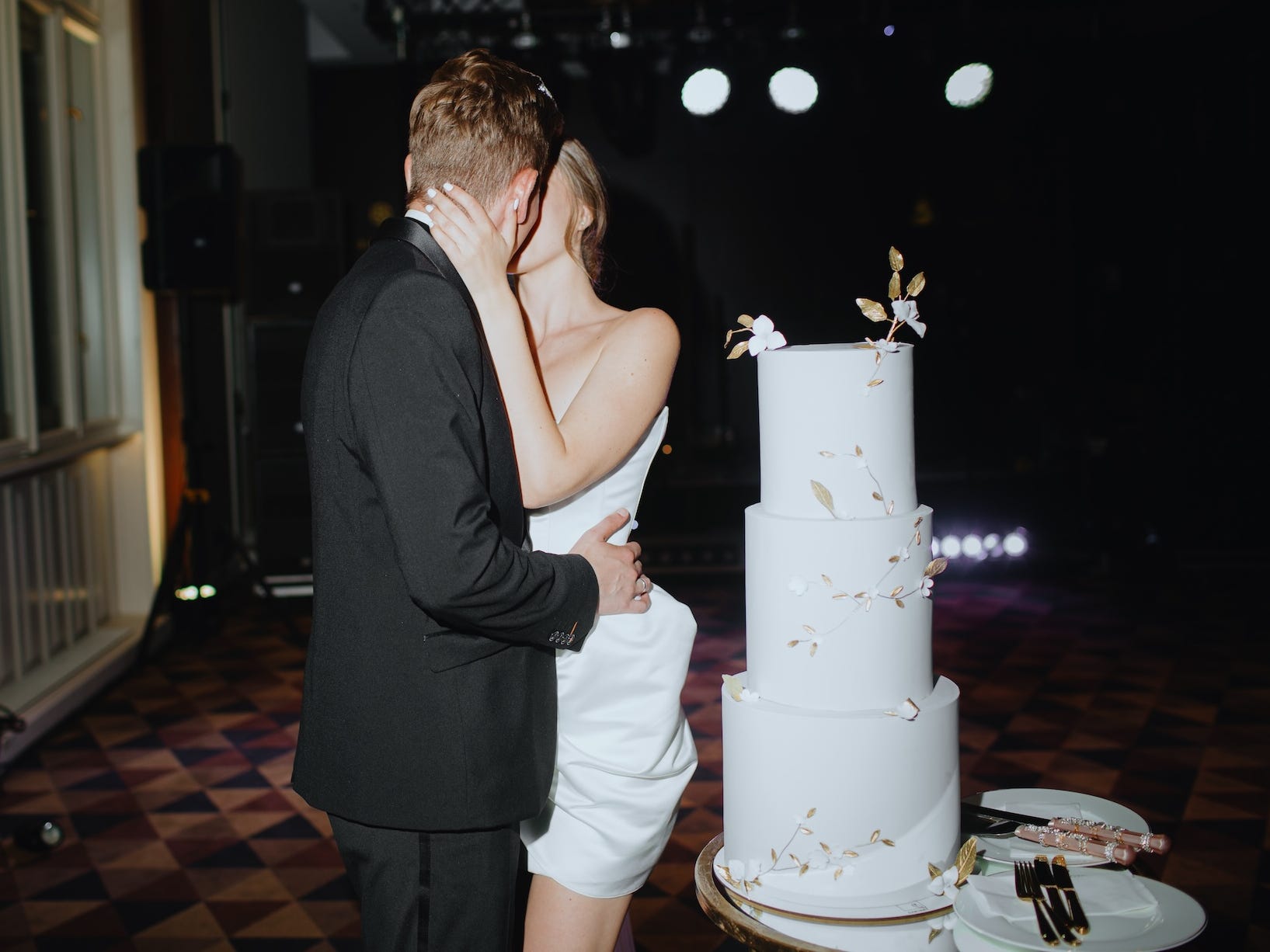 A couple kisses next to a three-tiered, white wedding cake.