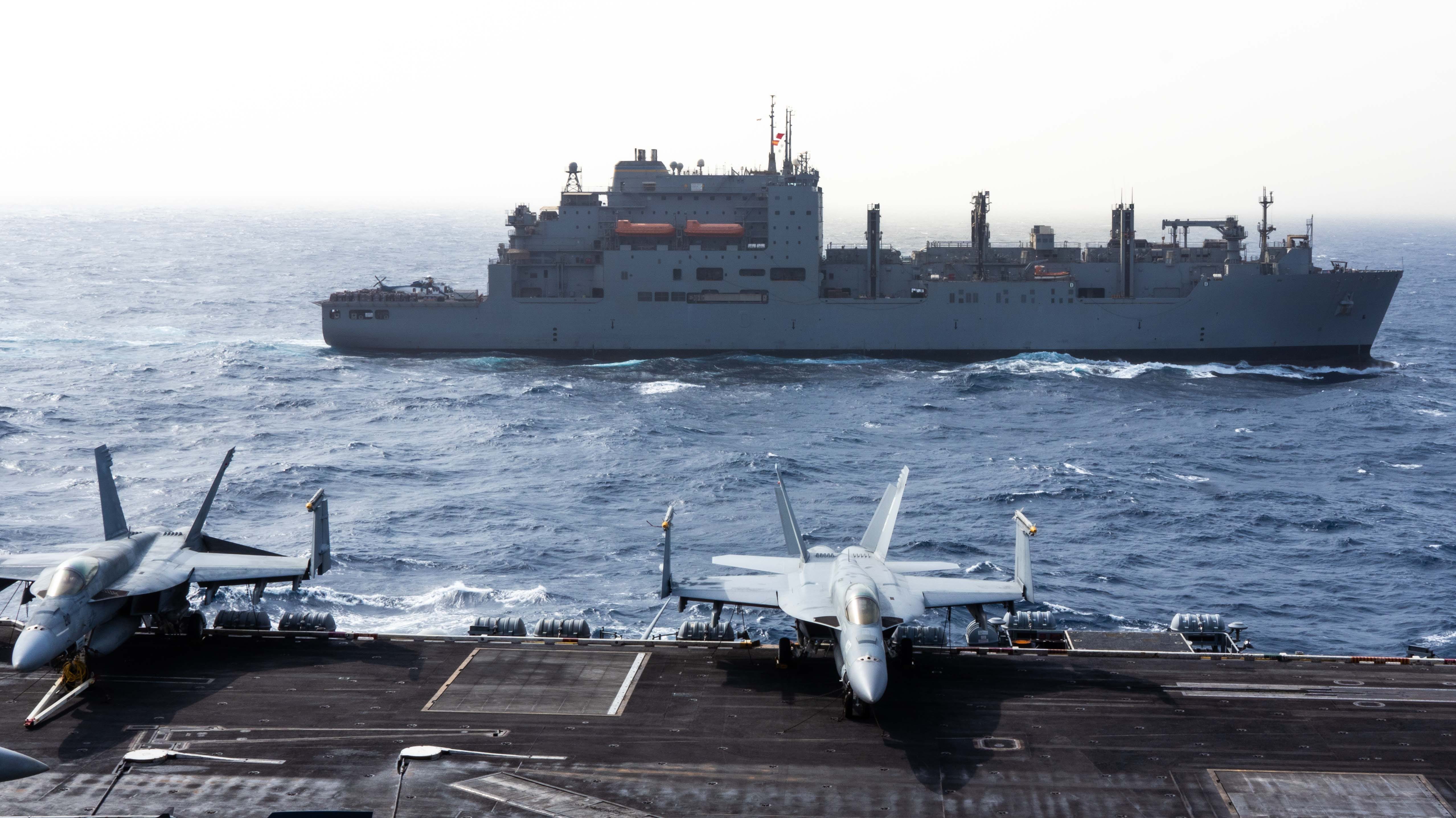 A cargo ship sails in blue waters against a white sky in the background. In the foreground, two aircraft sit on top of the tarmac of an aircraft carrier.