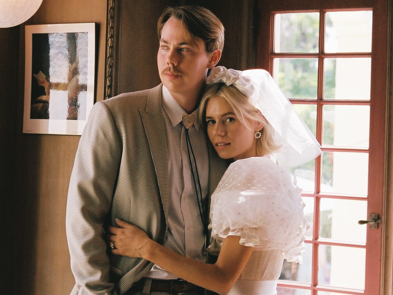 A bride and groom stand in a room embracing surrounded by windows.