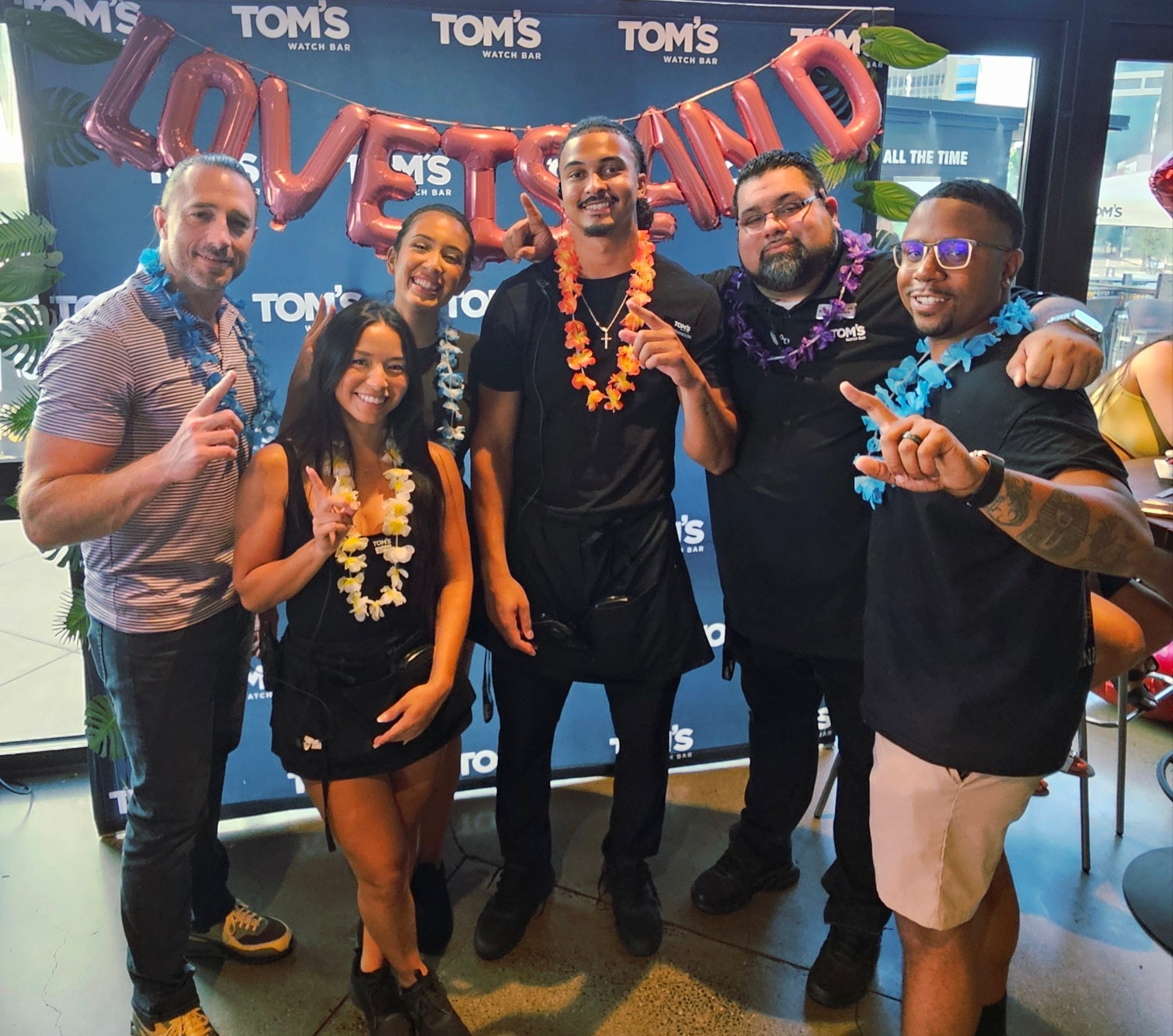 A group of people at Tom's Watch Bar pose in front of a 
