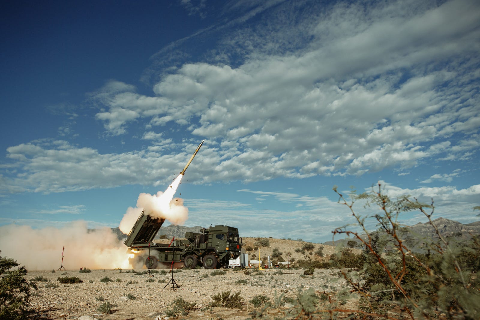 A missile is fired from a launcher on a track in the desert with a blue cloudy sky in the background.