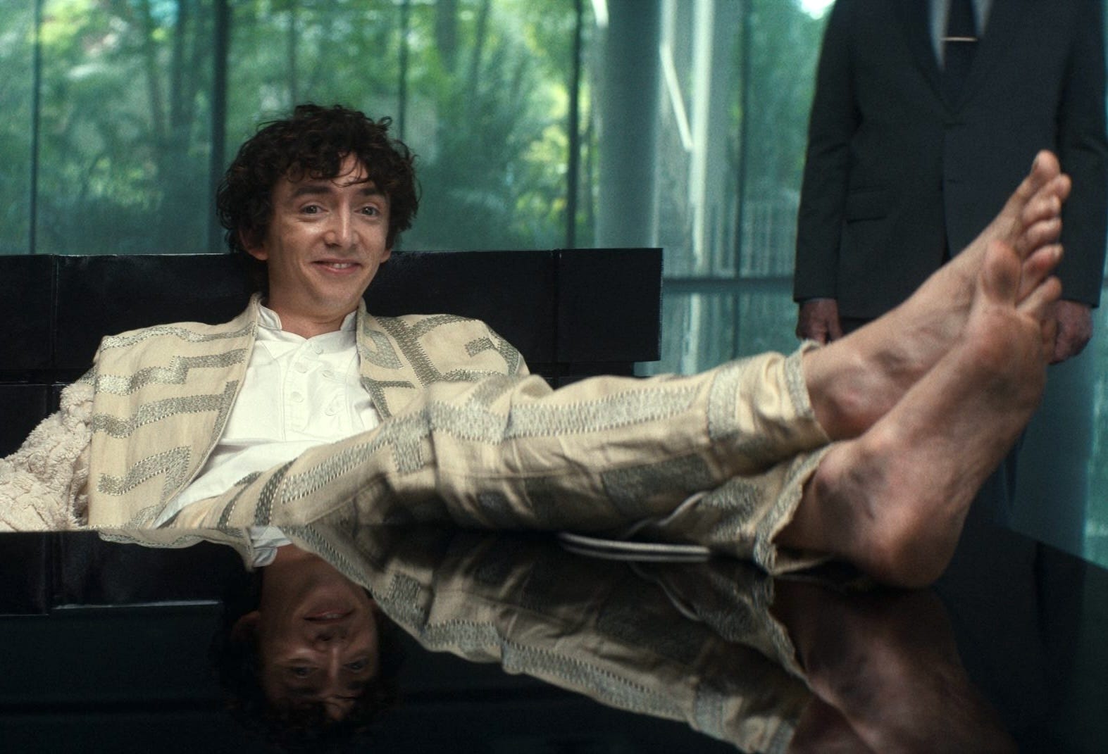 A young man with curly brown hair wears a beige and gold suit with a white shirt. He is resting his dirty bare feet up on a table.