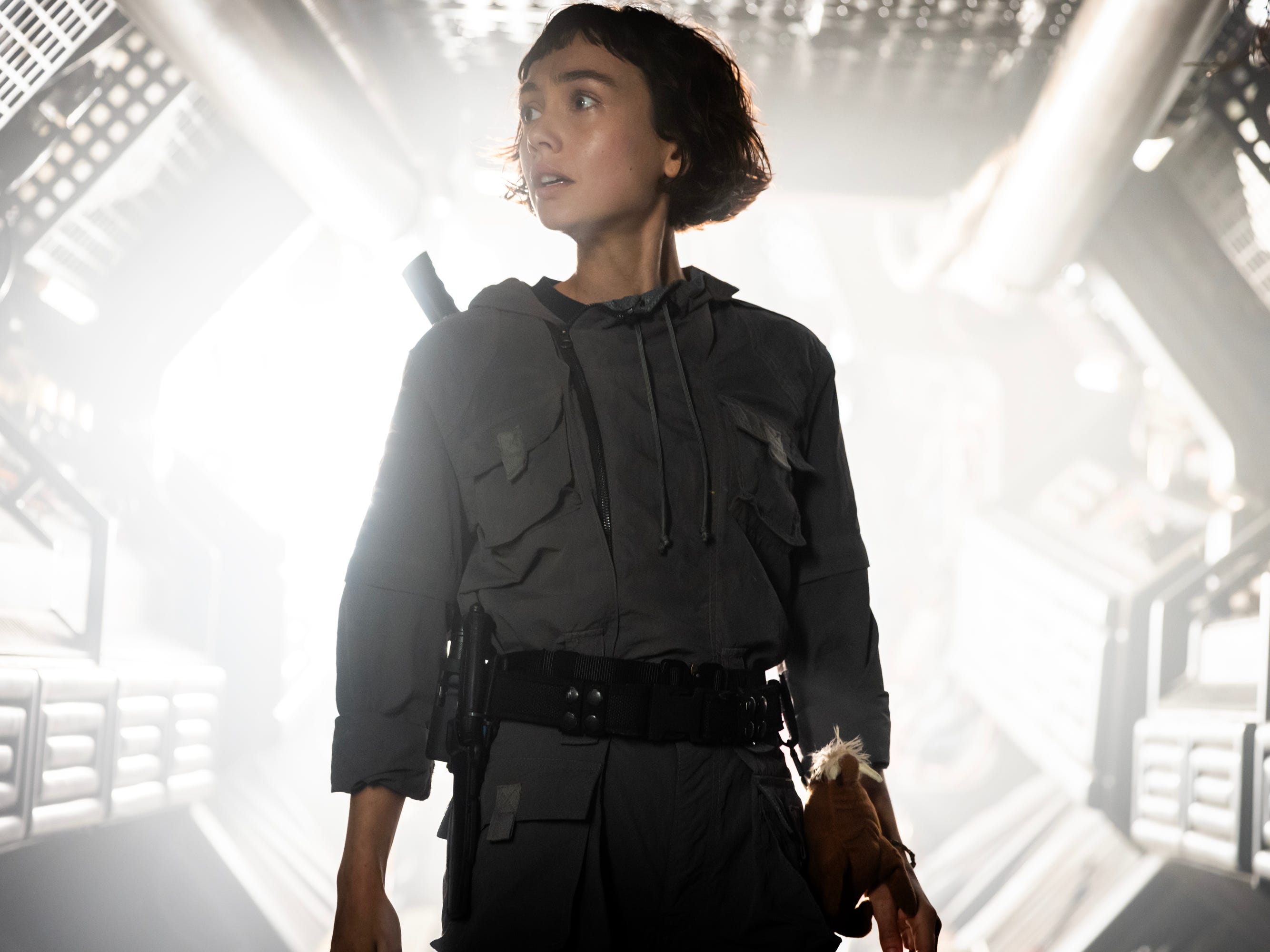A woman with short brown hair looks to the left. She's wearing a gray boiler suit and has a black belt around her waist. There is also a cuddly toy strapped to her side.