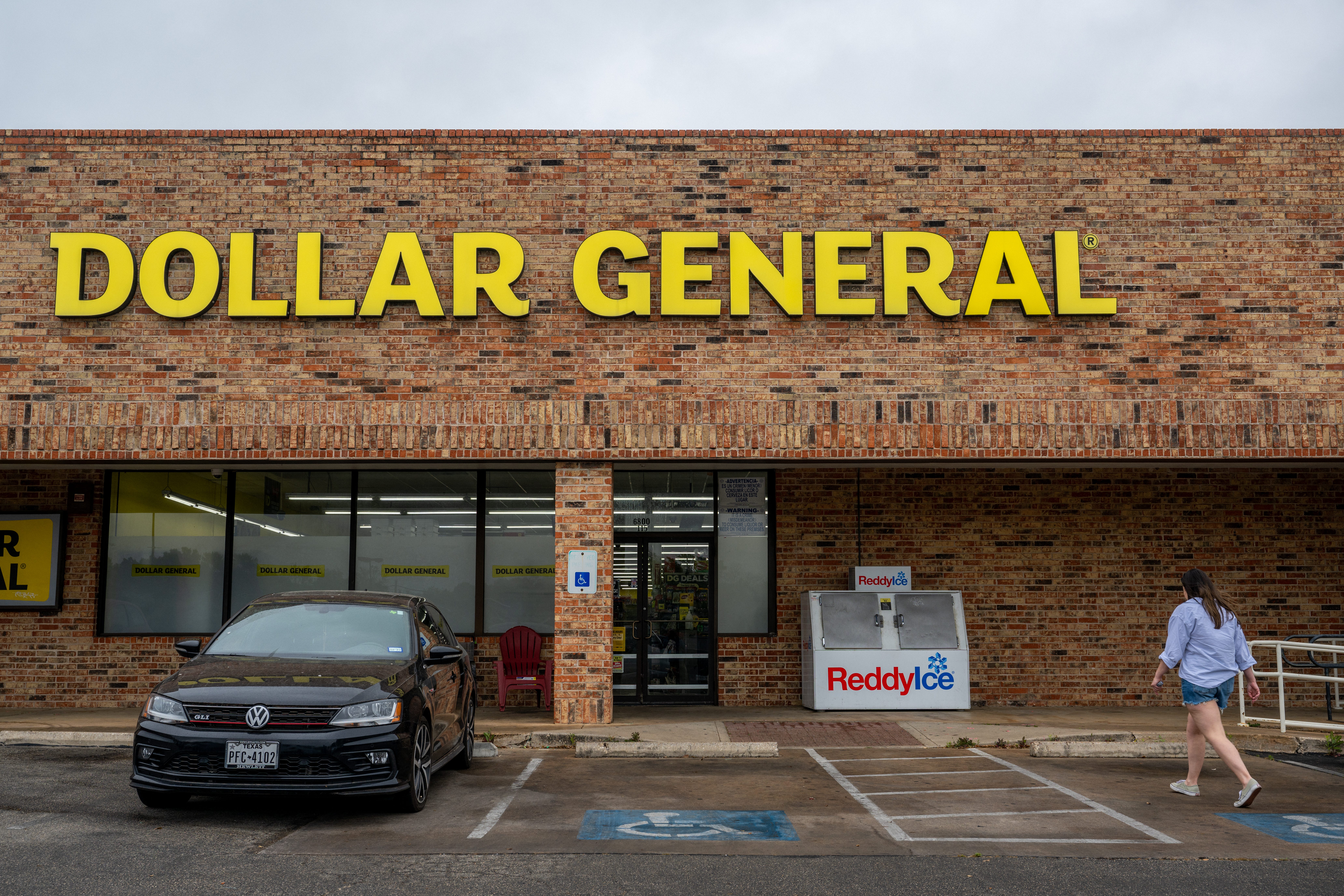 A customer walks into a Dollar General store though the parking lot in Austin, Texas. A compact Volkswagen car sits in front of the store, with the yellow "Dollar General" sign above the entrance and an overcast sky.