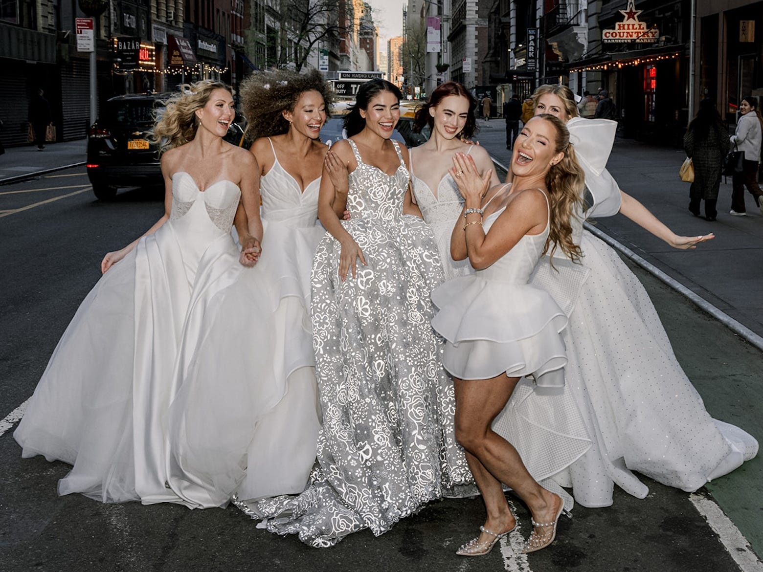 Hayley Paige claps in front of a group of women wearing wedding dresses on a street.