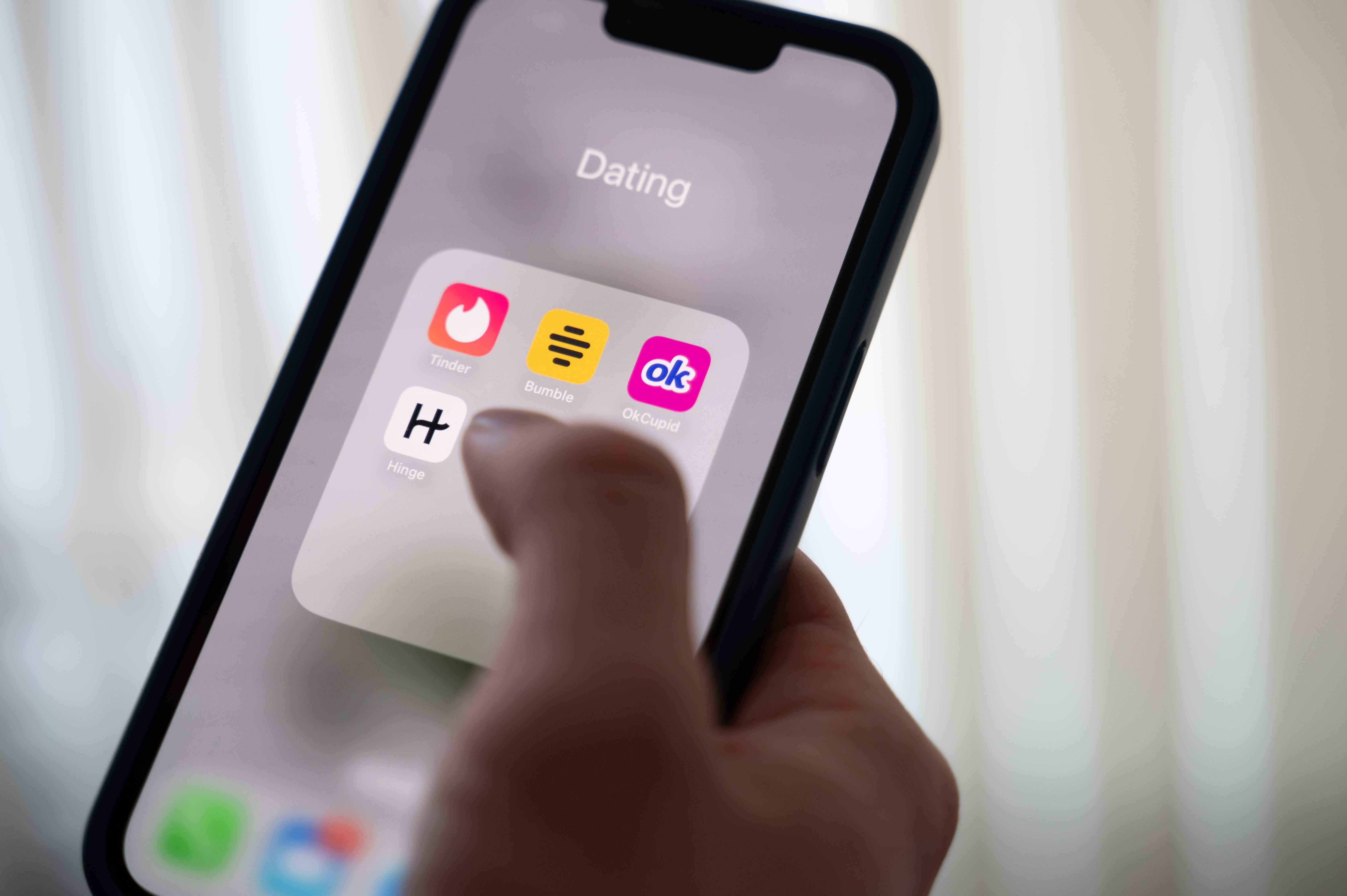 A hand hovers over an iPhone with dating app icons on display like Tinder, Hinge, and Bumble.