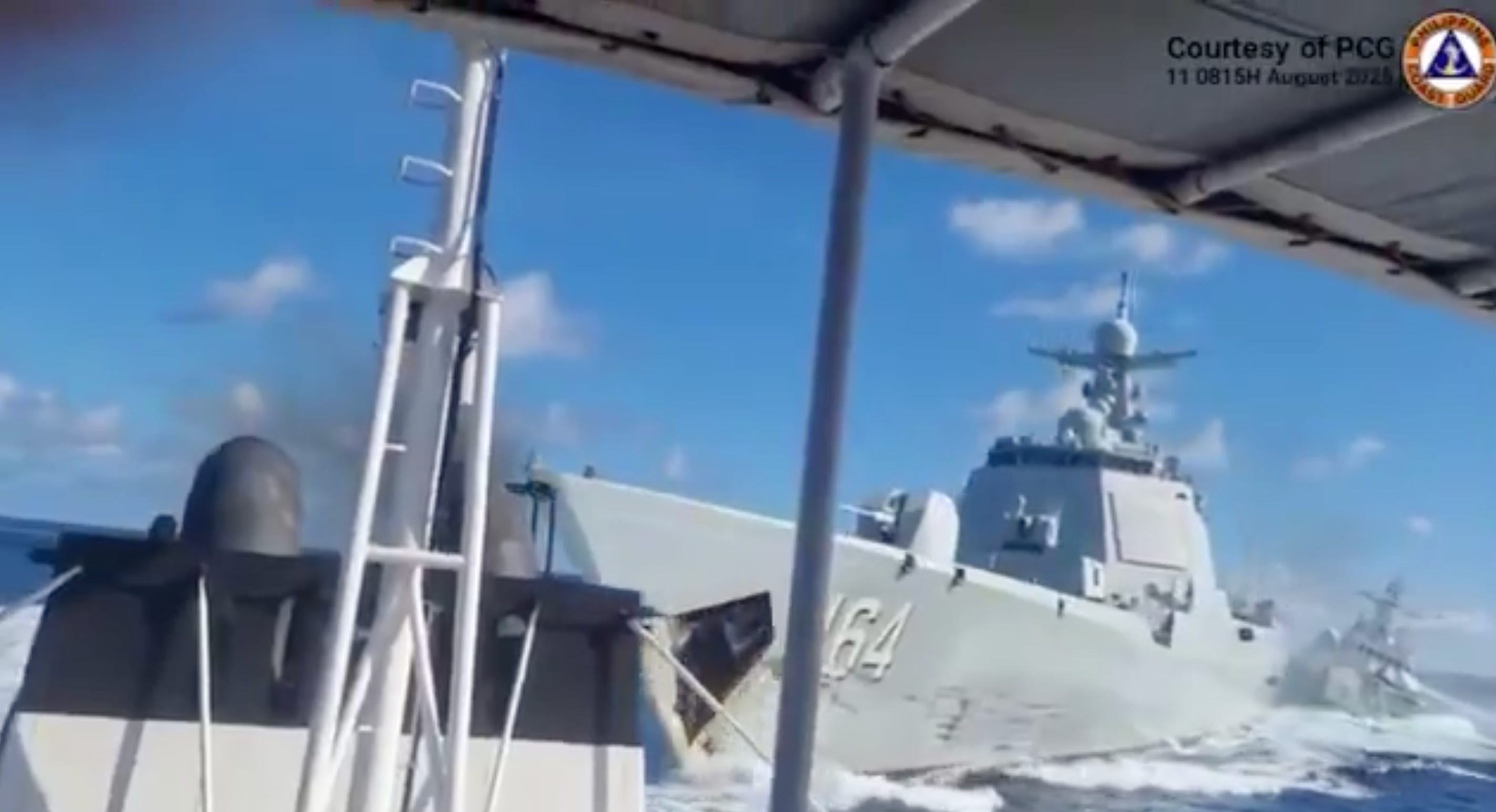 A screenshot of a video showing a Chinese Navy vessel and Chinese Coast Guard vessel crashing into one another in the sea. The video is filming from a fishing boat. The sky is blue with some clouds in the background.