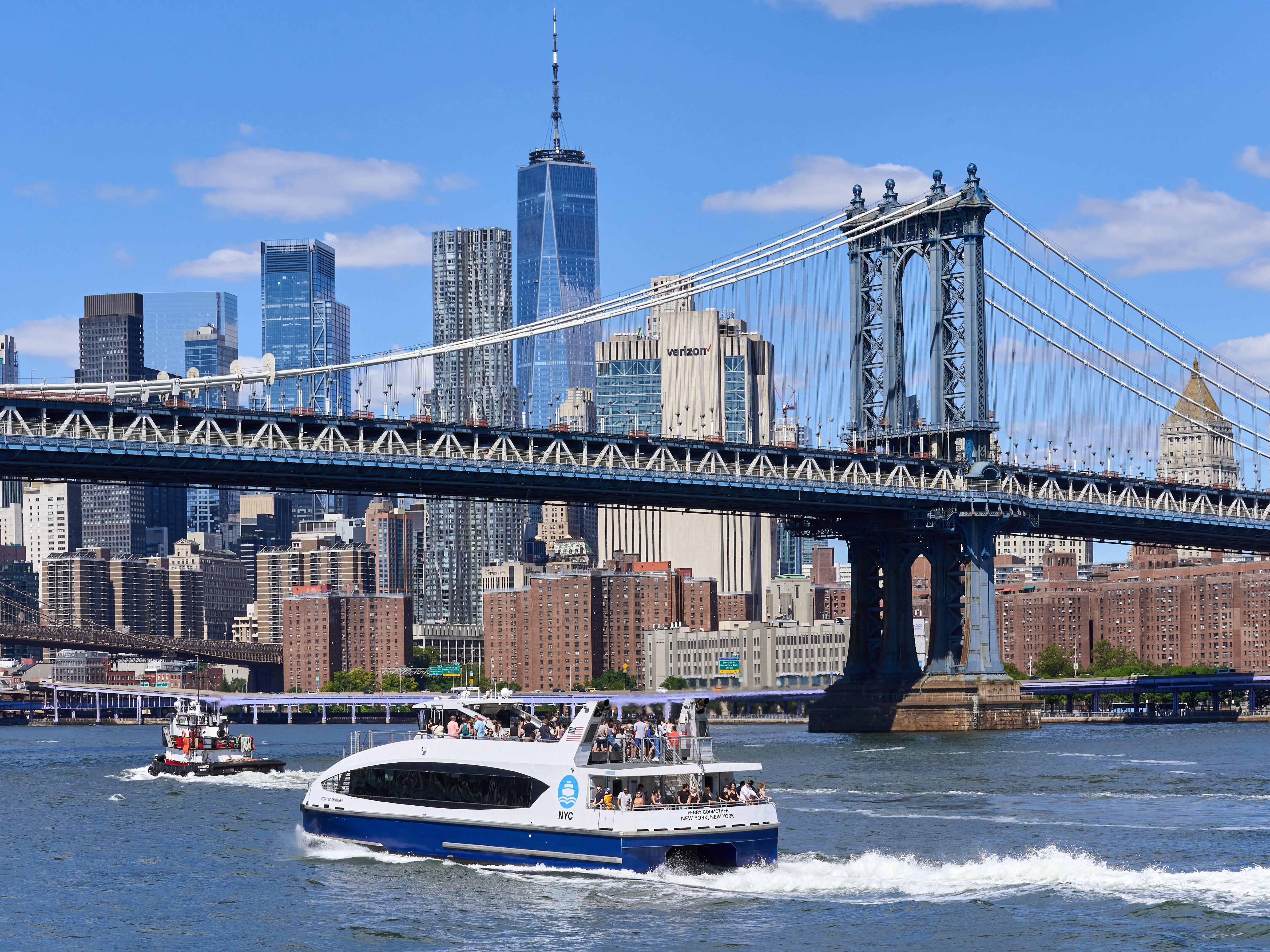 A NYC Ferry traveling on the East River with the Manhattan Bridge and skyscrapers in the background.