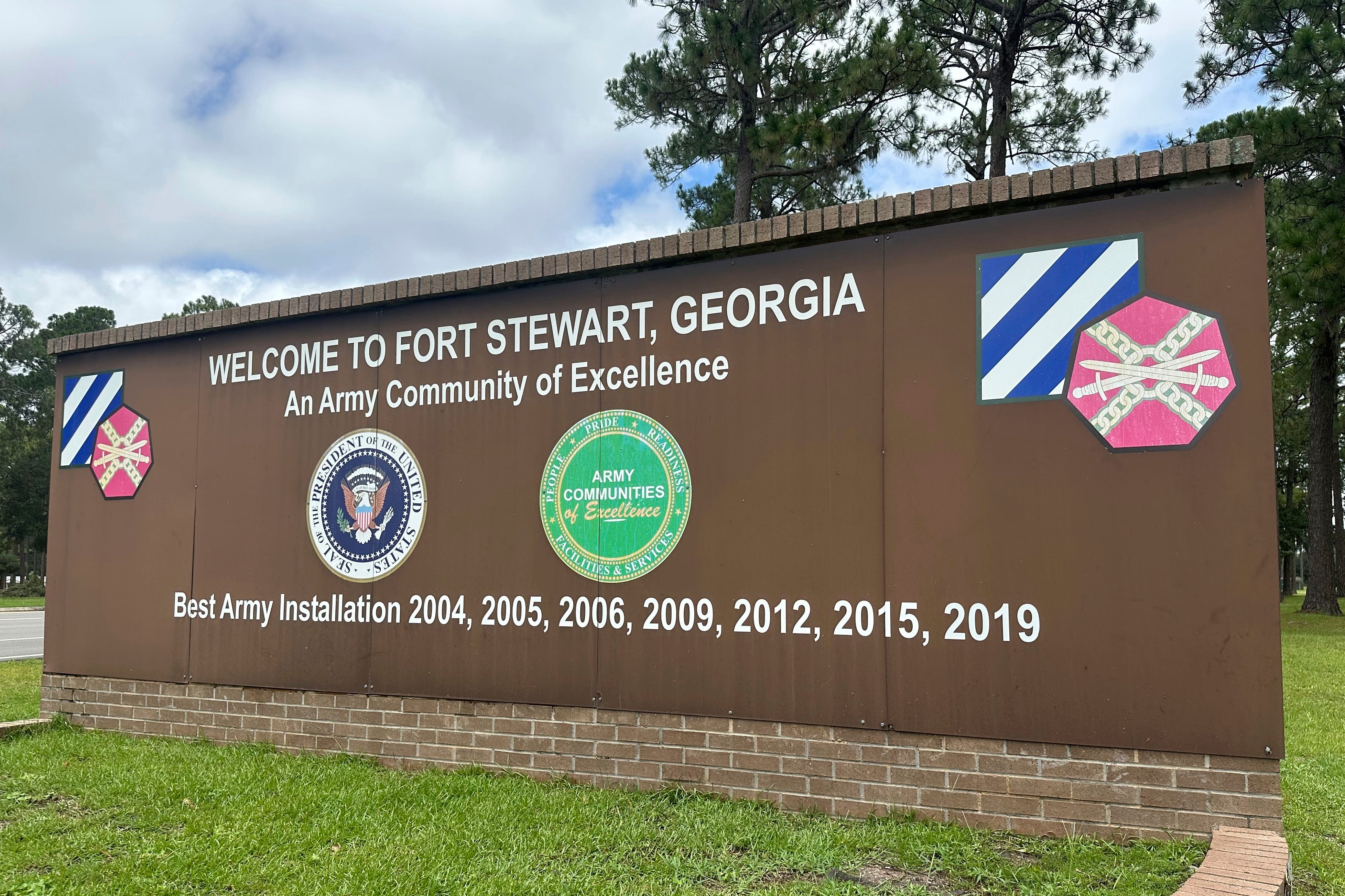 The gate sign of Fort Stewart, Georgia, is pictured. The grass is green in the foreground and the sky is cloudy in the background.