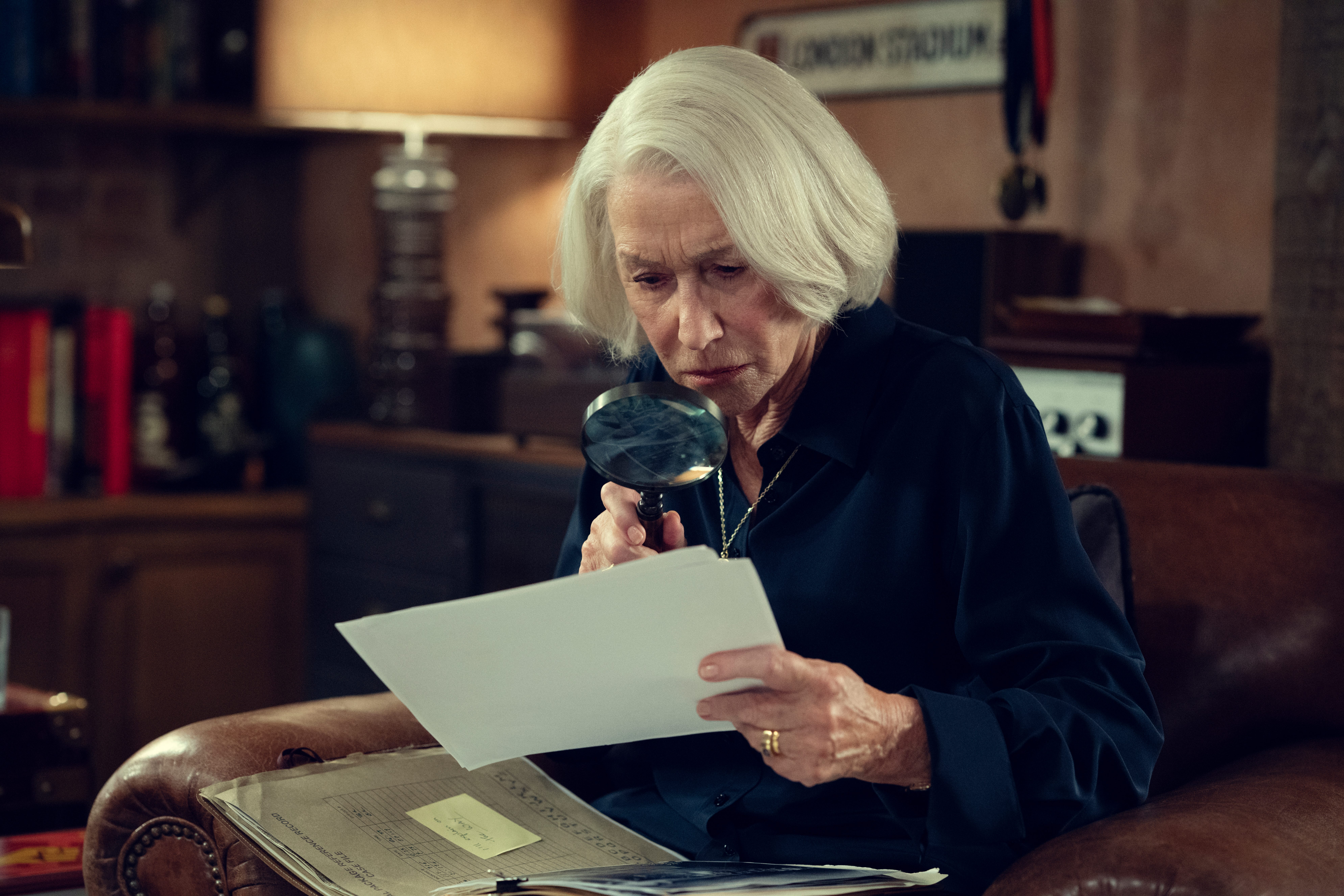 Helen Mirren holding a magnifying glass up against a document