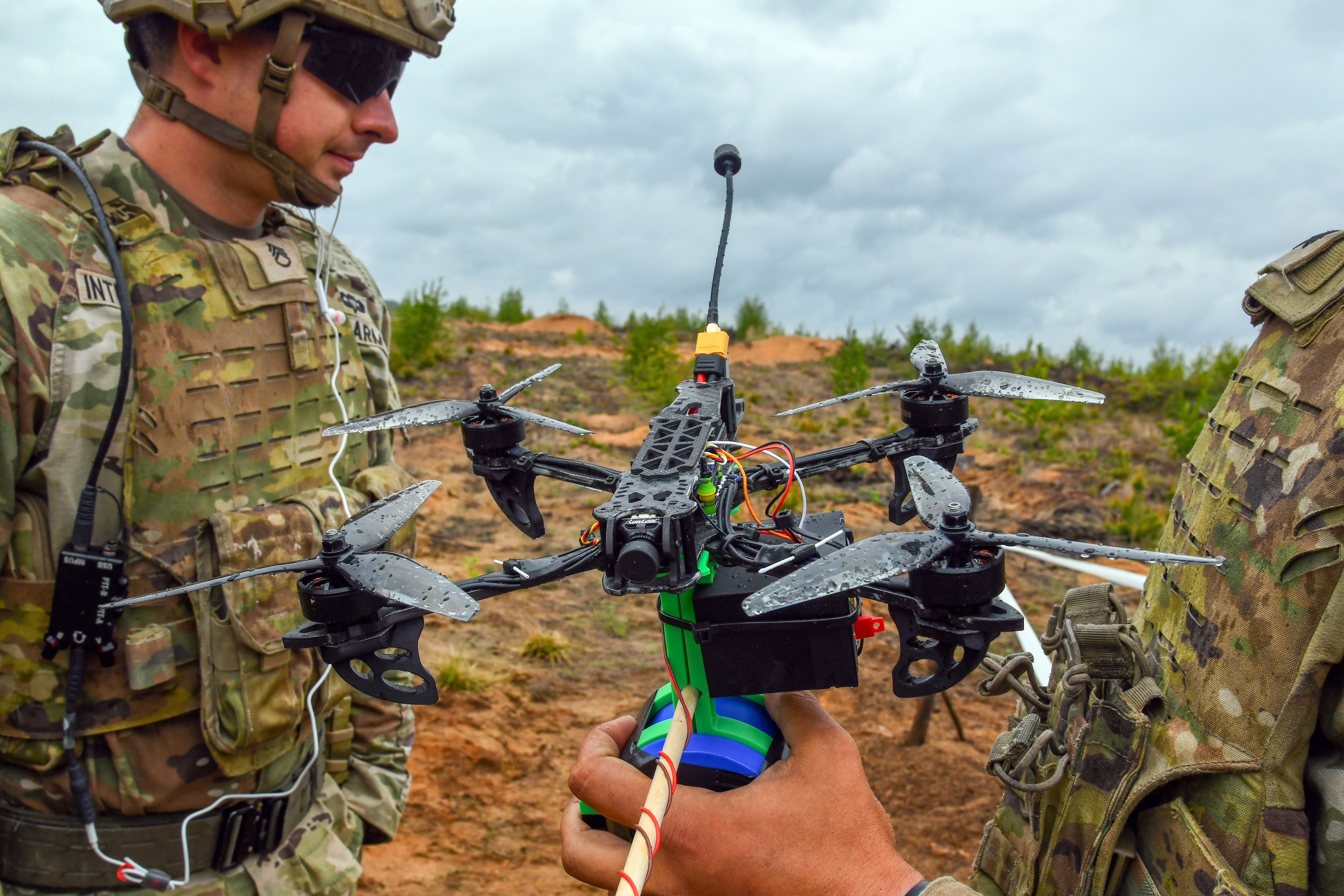 Two US Army soldiers look at a first-person view quadcopter drone on a muddy, grassy terrain with a cloudy sky in the background.