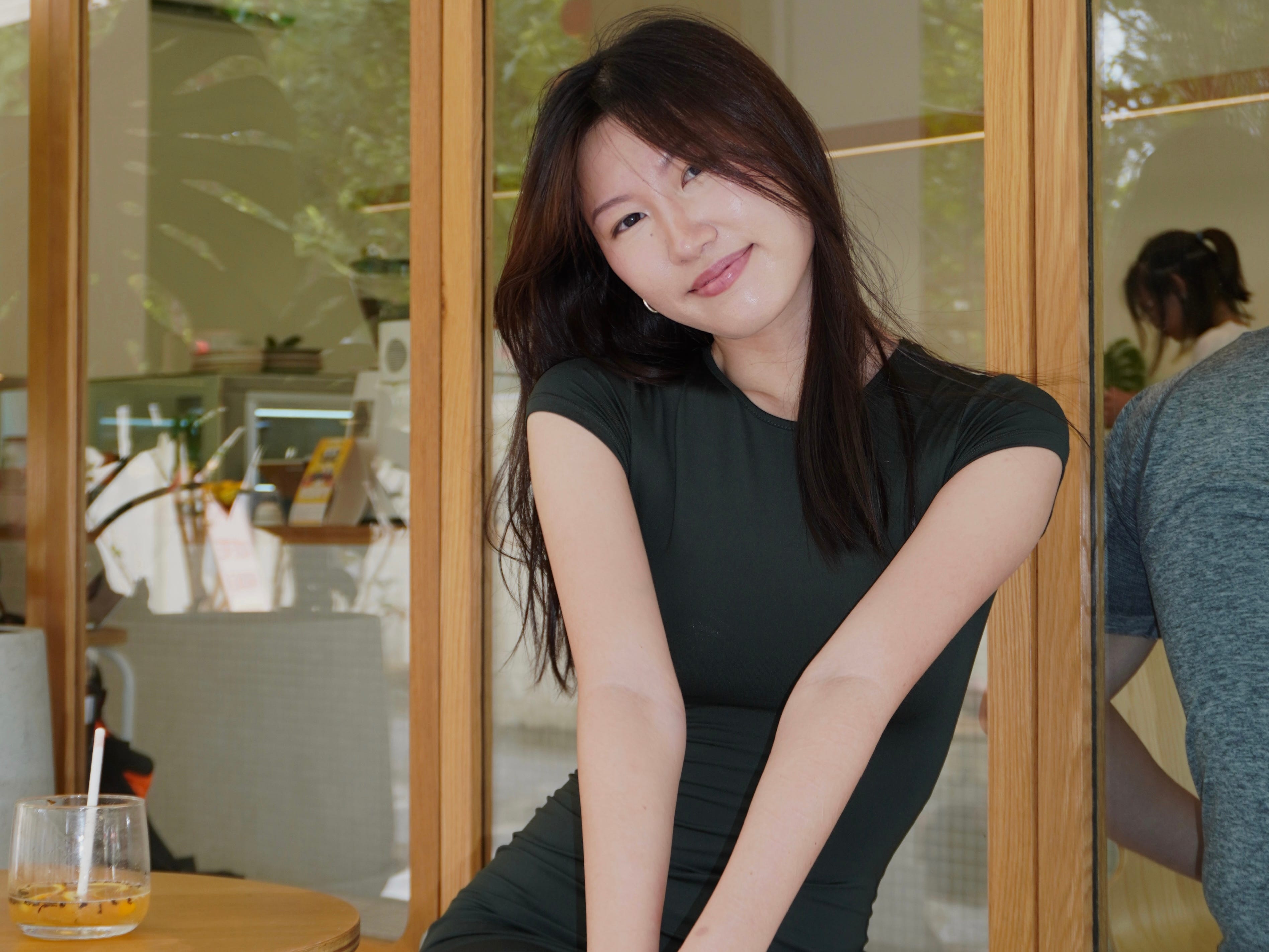 Evelyn Ramli sitting in front of a window wearing a black dress with a drink on the table by her side.