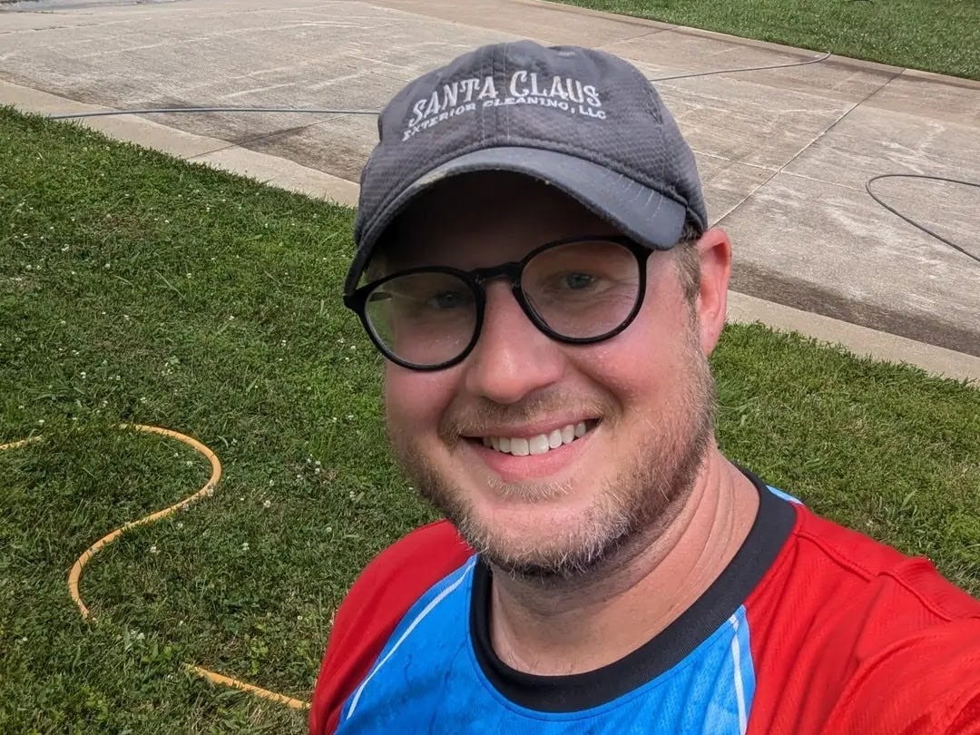 A man wearing glasses, a gray baseball cap, and a red and blue shirt smiles for a selfie. He is standing on a grassy lawn with a driveway and trailer in the background.