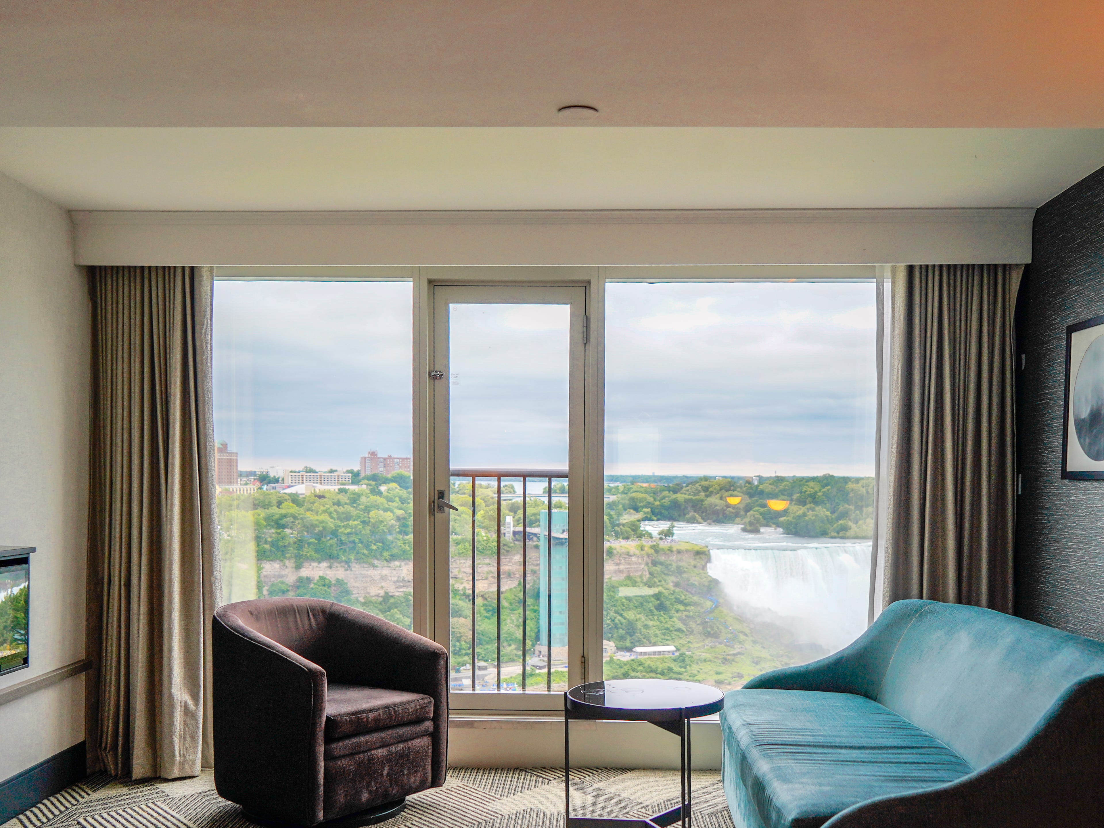 A table, sofa, and chair in a hotel room backed by floor-to-ceiling windows and a Juliette balcony facing Niagara Falls
