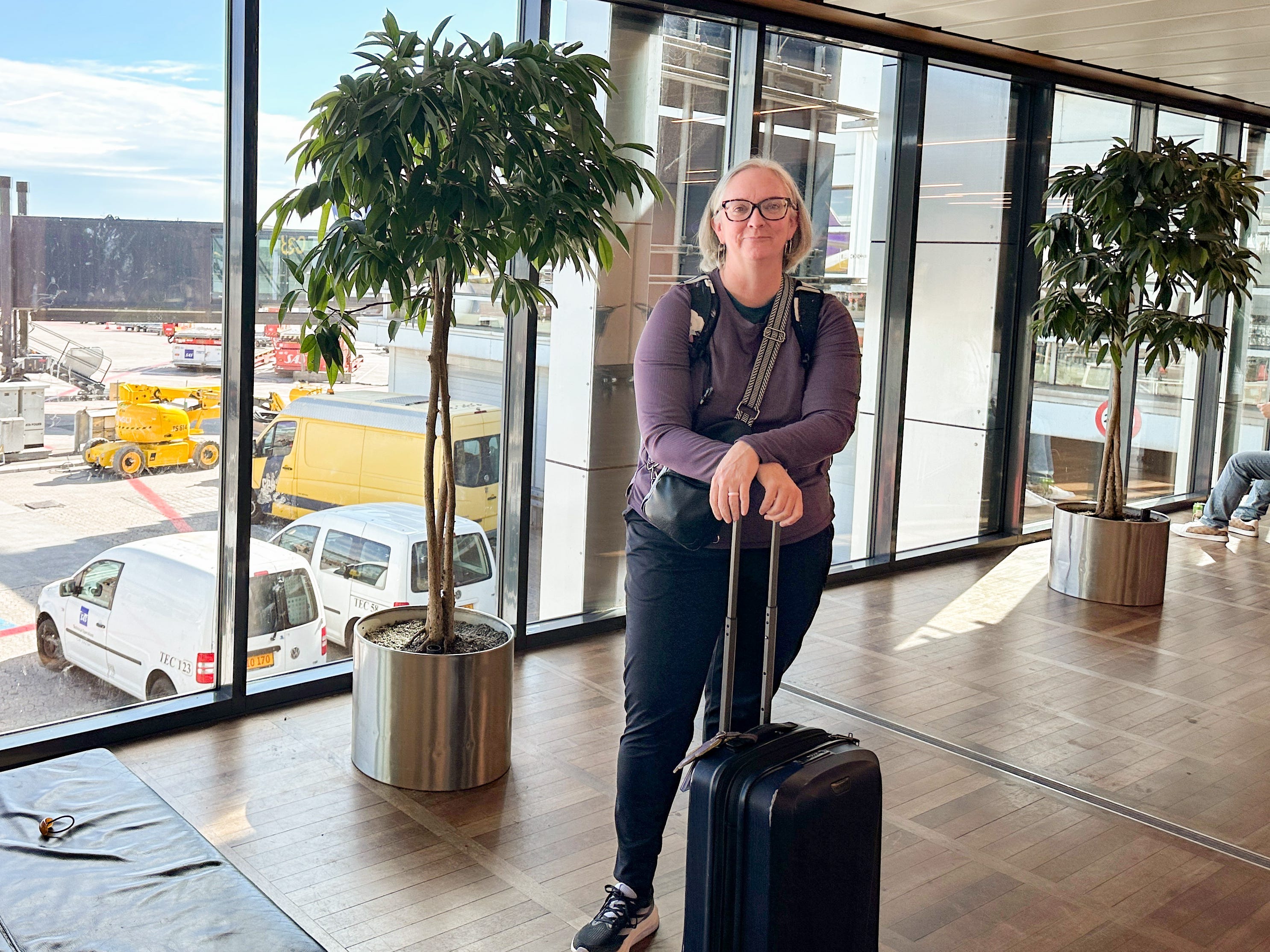 Kate stands in front of a window at the airport with a carry-on suitcase.