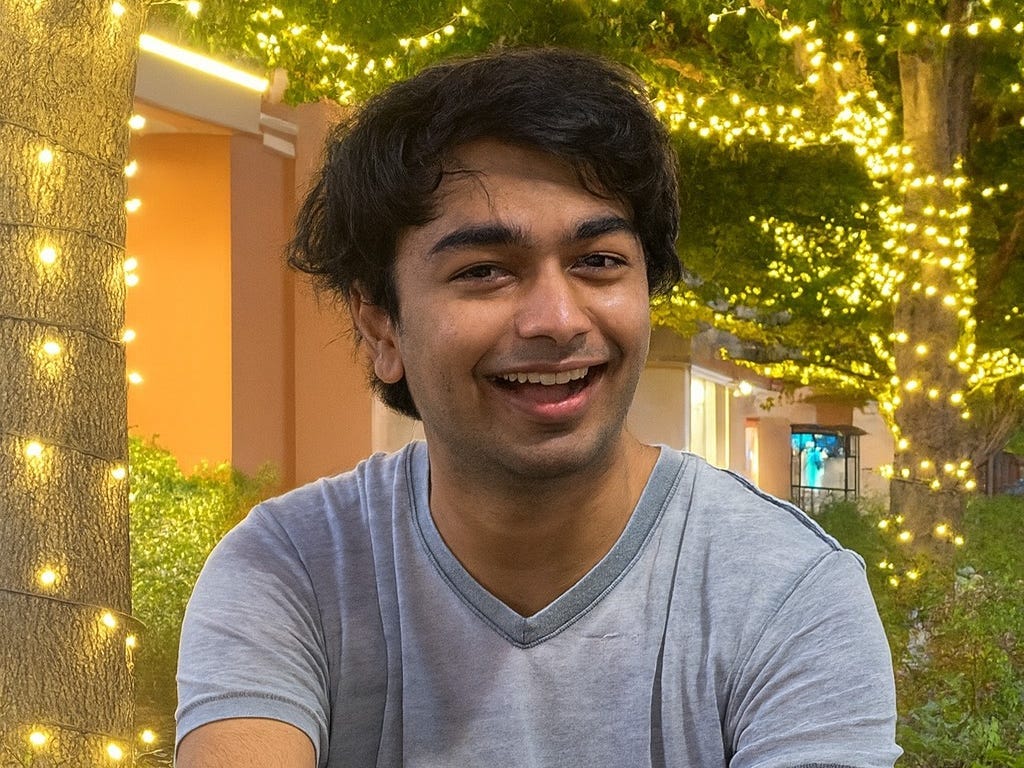 Smiling young person in a gray t-shirt, seated outdoors with blurred string lights in the background.