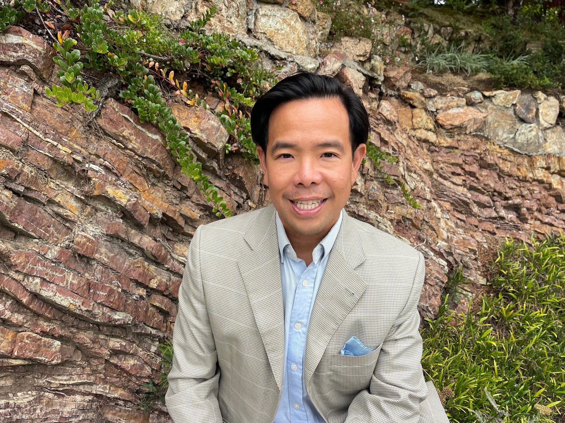 Smiling man in a light blazer with a blue shirt and pocket square, seated outdoors against a rock wall with greenery.