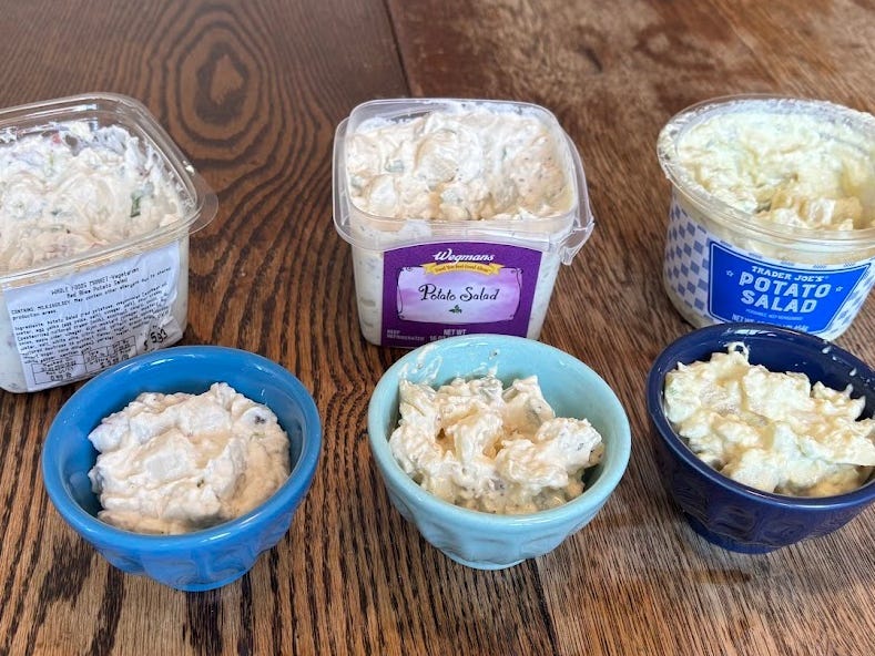 Containers of Whole Foods Red Bliss potato salad, Wegmans potato salad, and Trader Joe's potato salad, each with a small bowl in front displaying the corresponding salad.
