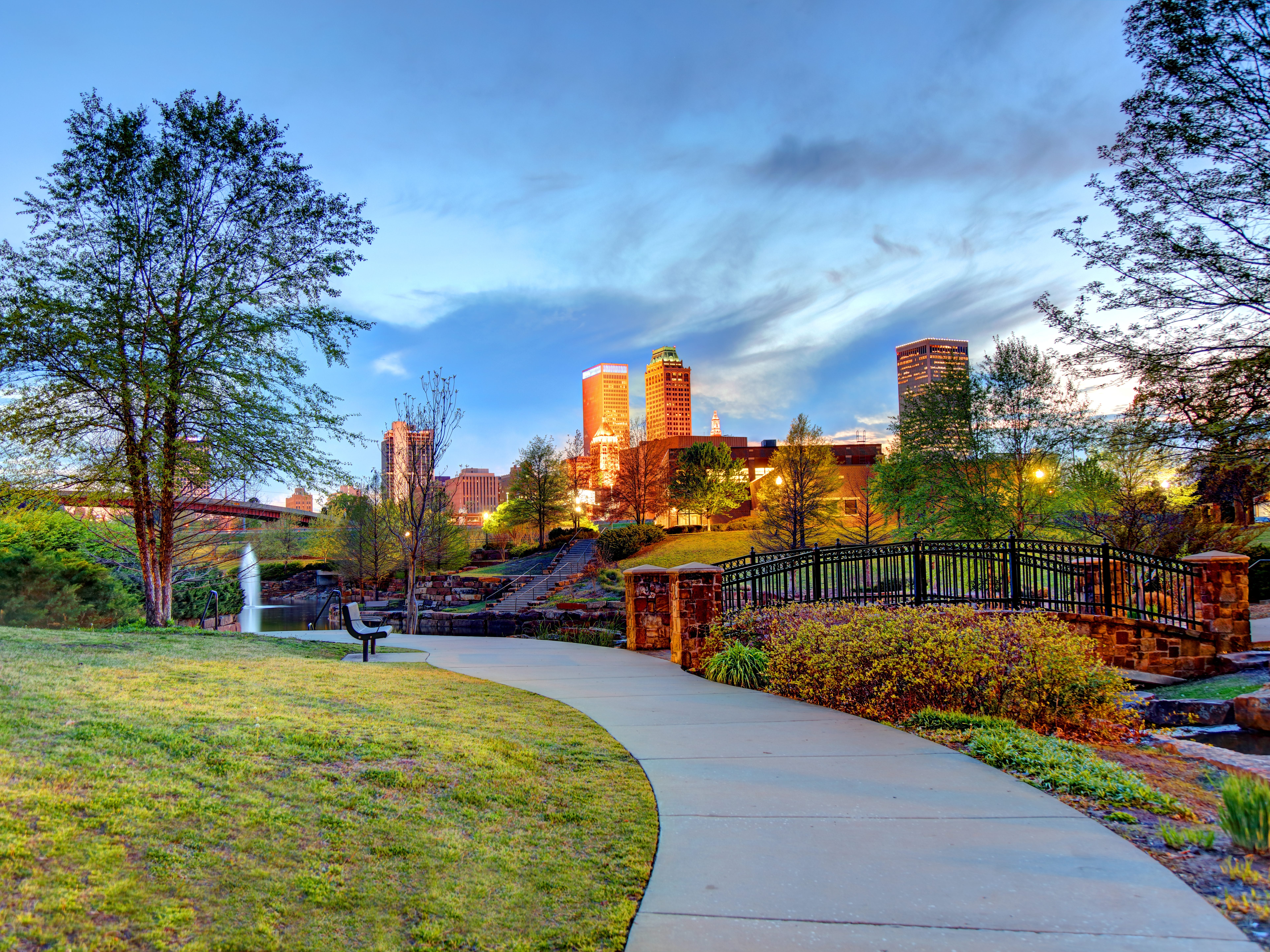 A park with a bridge, trees, and the Tulsa skyline in the background.