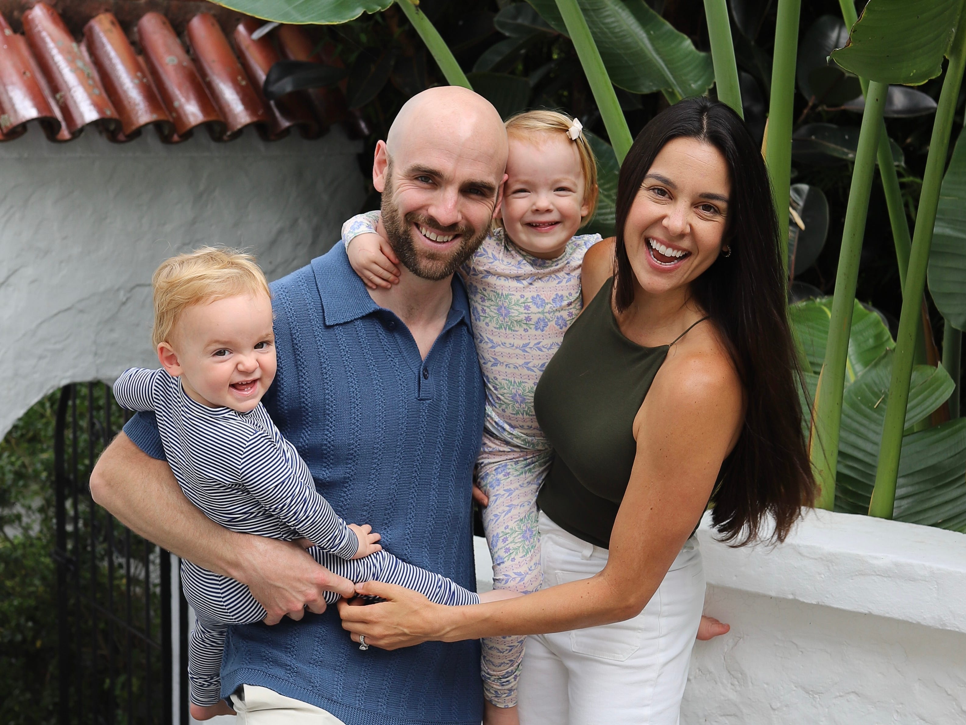 Samantha Gold with her family in Sydney. They are outside in front of a birds-of-paradise plant, all smiling, and the children are wearing printed pajamas.