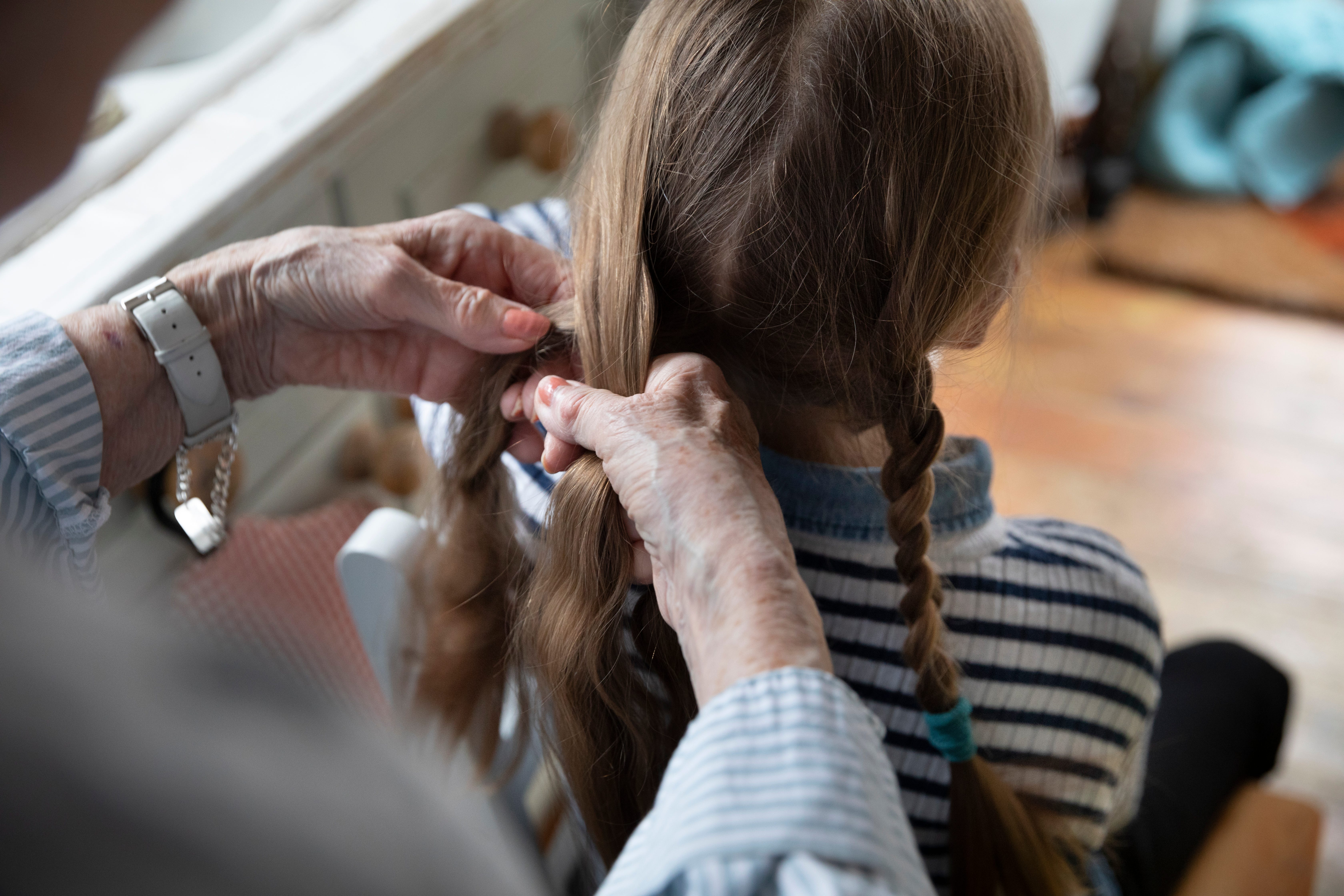 Over the shoulder view of Grandmother's hands, braiding a little girl's brown hair in a domestic setting.