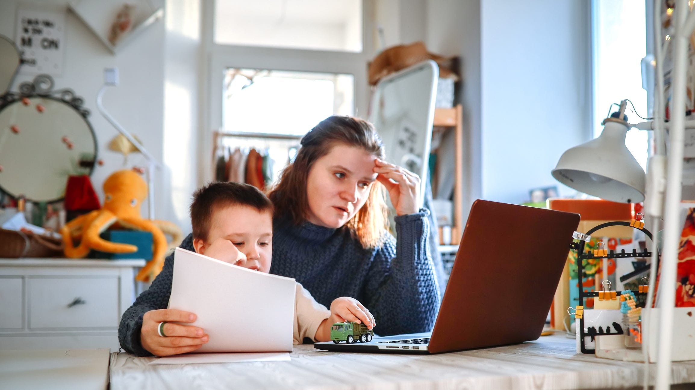 A stock photo of a tired woman with a toddler in her arms as she looks at a computer